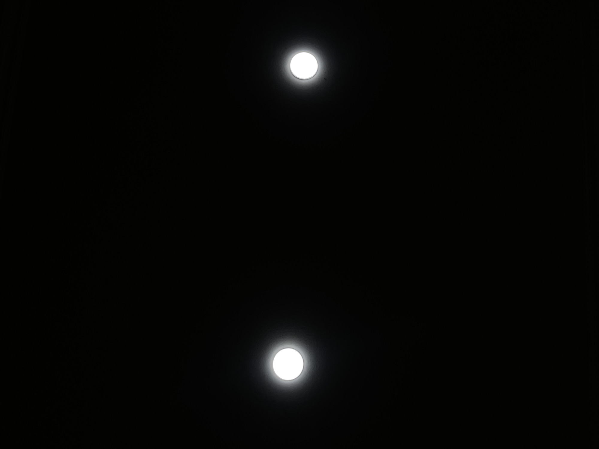 The image is a vertical, full-frame photograph of a dark, possibly night, environment. Two bright, circular light sources are visible, each with a faint halo. The light on the left is positioned higher and more to the left of the frame, while the light on the right is lower and more to the right. The background is uniformly black, suggesting the absence of any discernible features or environmental context. The image does not provide any clues or information related to the city of Tbilisi, Georgia.