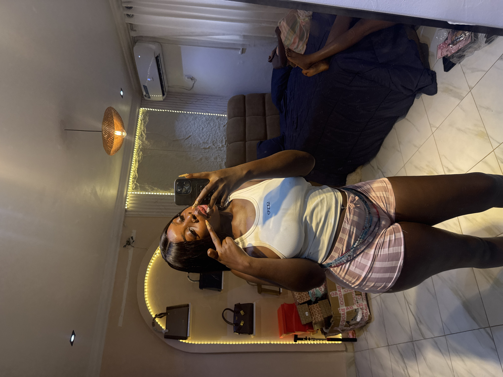 A young Black woman with dark hair and dark skin is taking a mirror selfie in a bedroom in Abuja, Nigeria. She is wearing a white tank top and pink and white striped shorts. She is making a playful gesture with her fingers near her mouth. The room is lit by warm yellow string lights and a pendant lamp. In the background, there is a bed with a dark blue comforter, an air conditioning unit, and shelves displaying handbags. The floor is tiled with a marble pattern.
