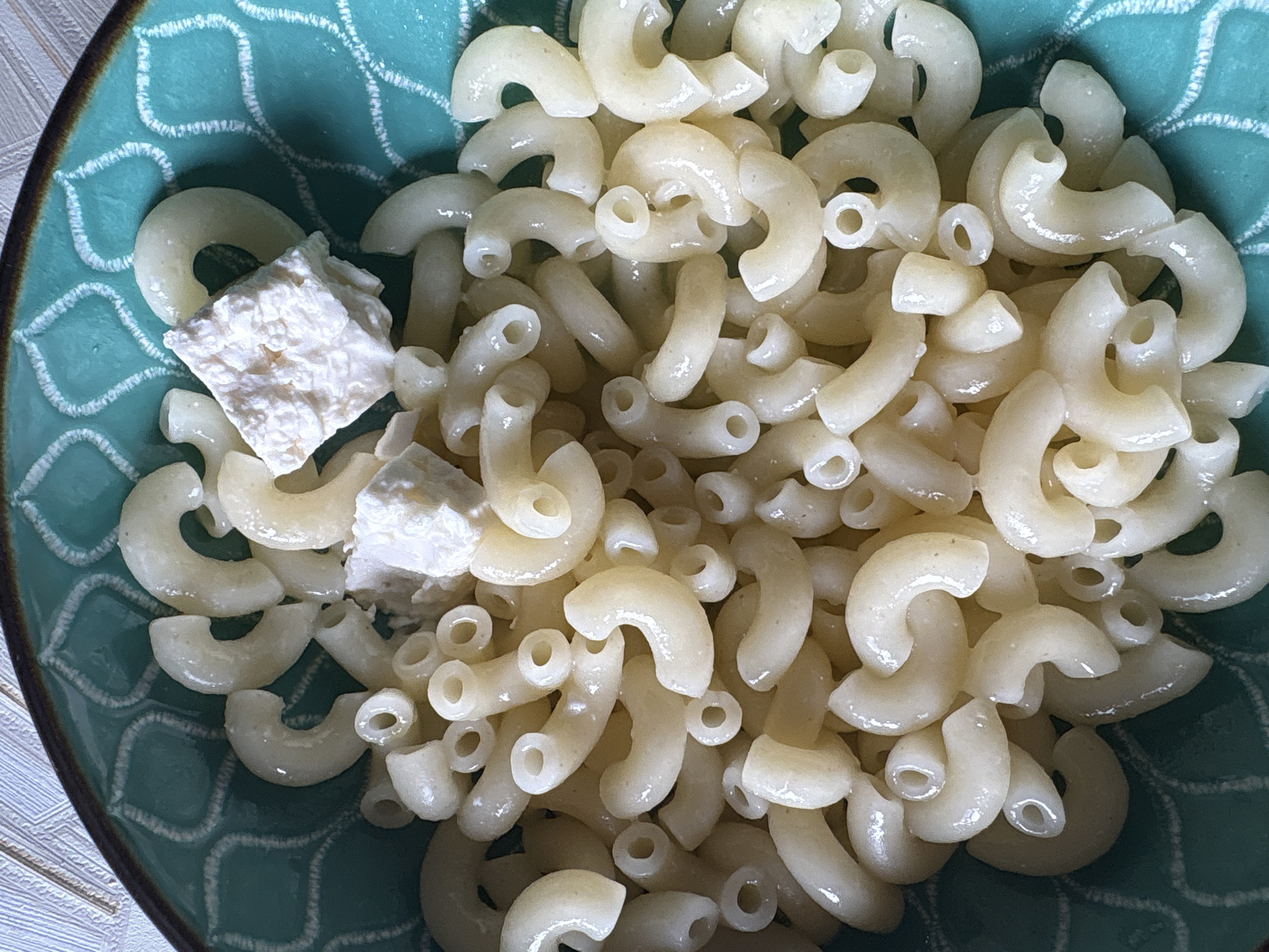 A close-up overhead shot shows elbow macaroni and crumbled feta cheese in a teal-colored bowl with a decorative pattern. The bowl is placed on a textured surface. The macaroni is cooked and appears slightly oily. Two pieces of feta cheese are visible, one larger than the other, and both are white and crumbly. The image appears to be taken indoors. There is no information about the location of Cheshnegirovo, Bulgaria, in this image.