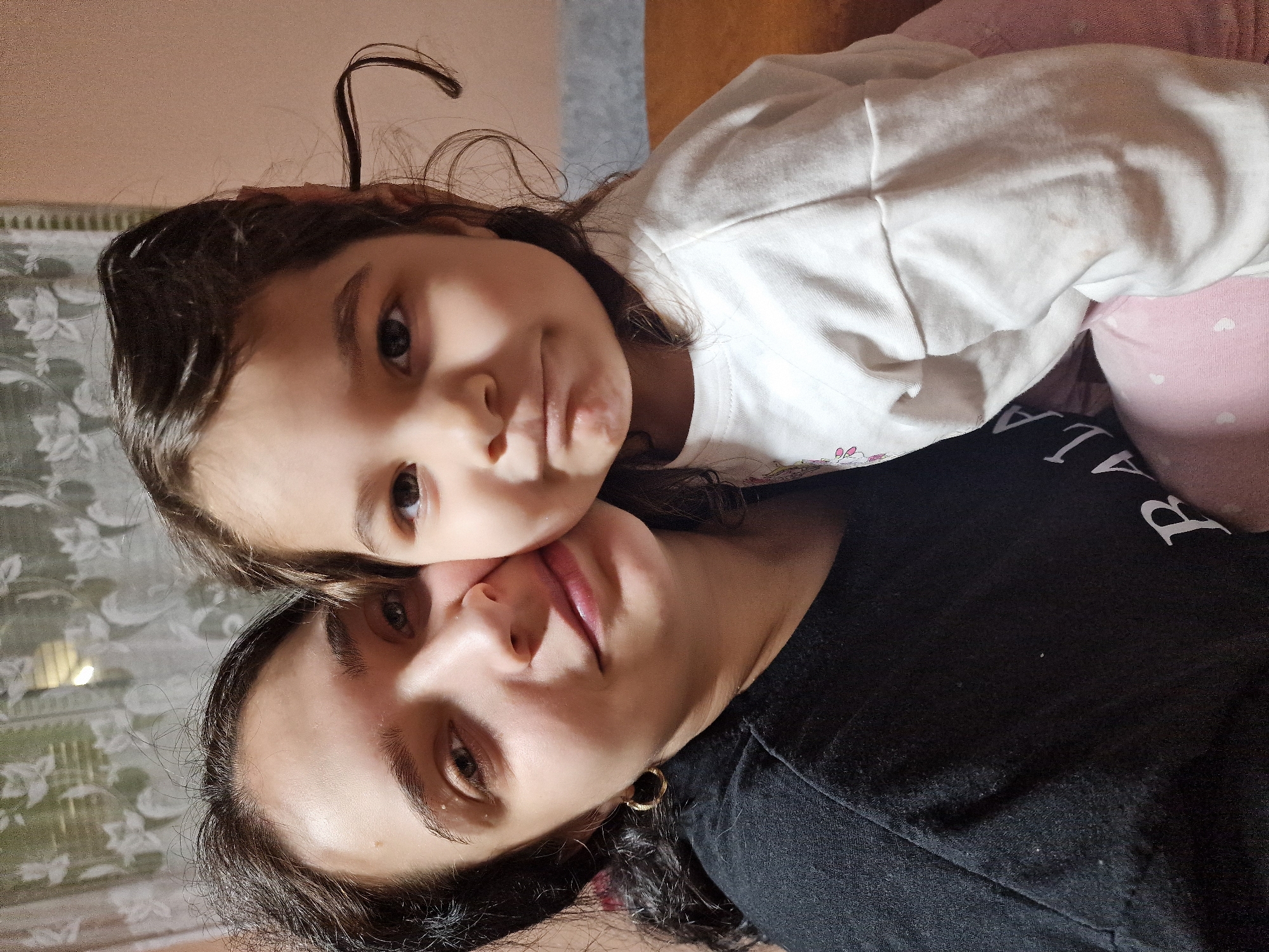 A close-up shot depicts a mother and child lying together. The child, positioned above, wears a white long-sleeved shirt. The mother, below, is dressed in a black shirt with white letters. The background is a blurry mix of pale pink, white and grey surfaces. The location appears to be indoors.