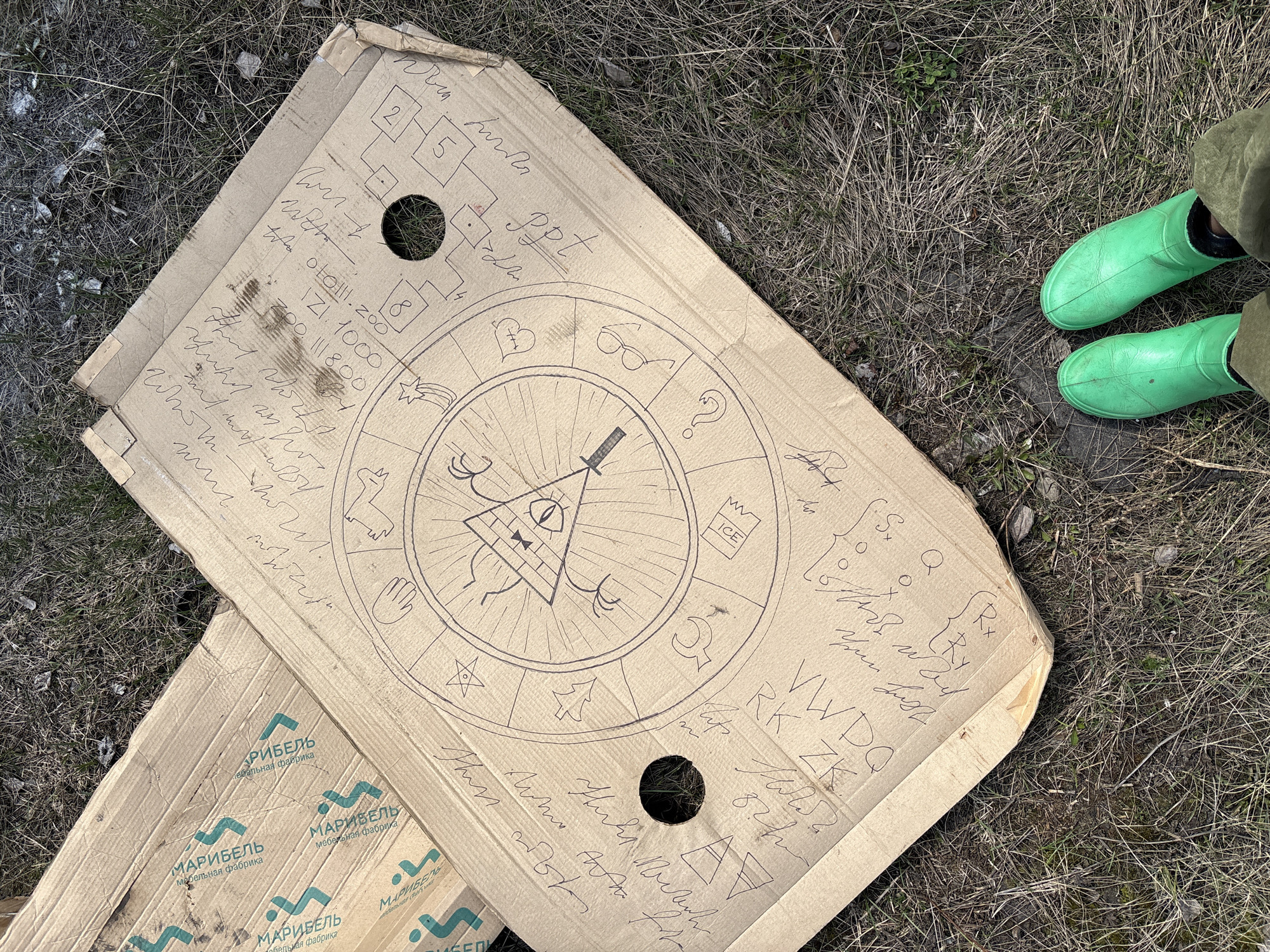 Here is a description of the image.
The image shows two cardboard boxes lying on dry grass. A pair of green rubber boots are visible on the right side of the image, partially obscured by the grass. The boxes are marked with the name "МАРИБЕЛЬ" (MARIBEL) in Cyrillic, followed by "мебельная фабрика" (furniture factory). The upper box is covered with doodles and writing in pen. At the center of this box, there is a drawing reminiscent of Bill Cipher from the show "Gravity Falls". There are also what looks like mathematical and scientific notations, along with strings of numbers and letters. Two holes have been cut into the cardboard. The image was most likely taken outdoors. I cannot confirm the location is the city of Zheleznodorozhny, Russia.
