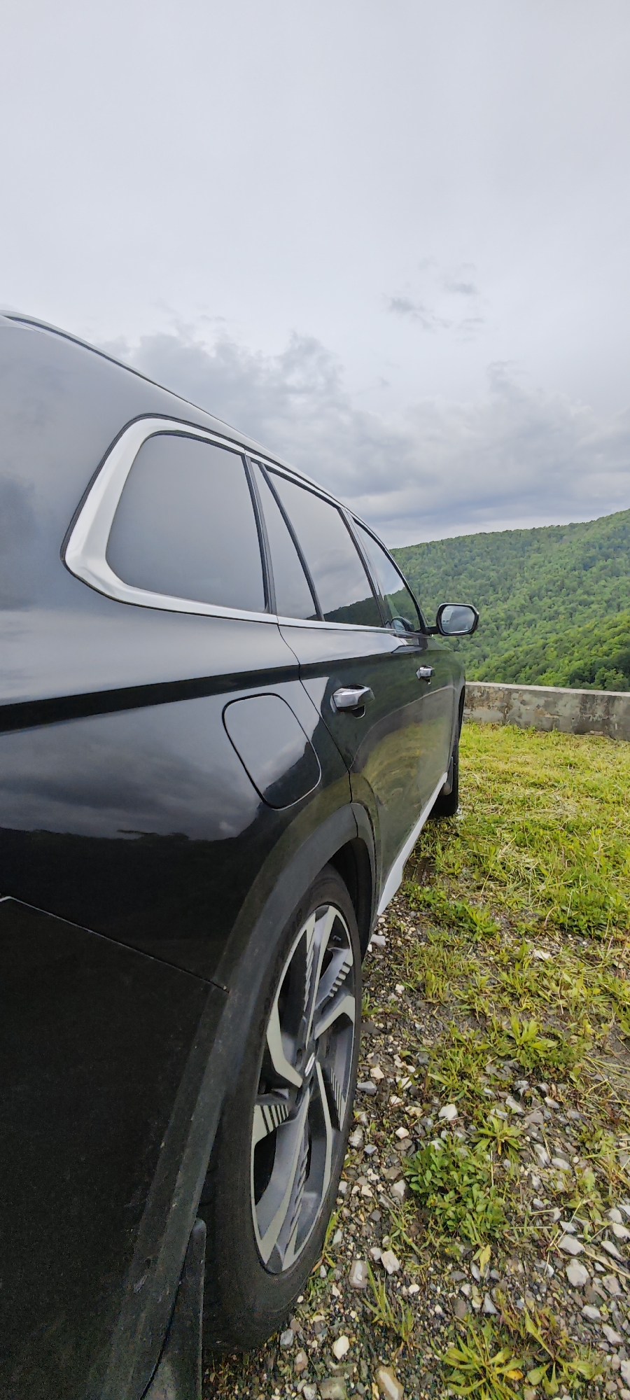 The image shows a black Hyundai Tucson parked on a gravel and grassy area near a concrete barrier. In the background, there are green forested hills and a cloudy sky. The location is Estesadok, Russia. The focus is on the rear of the vehicle, showing the wheel well, tire, and window. No people are visible.