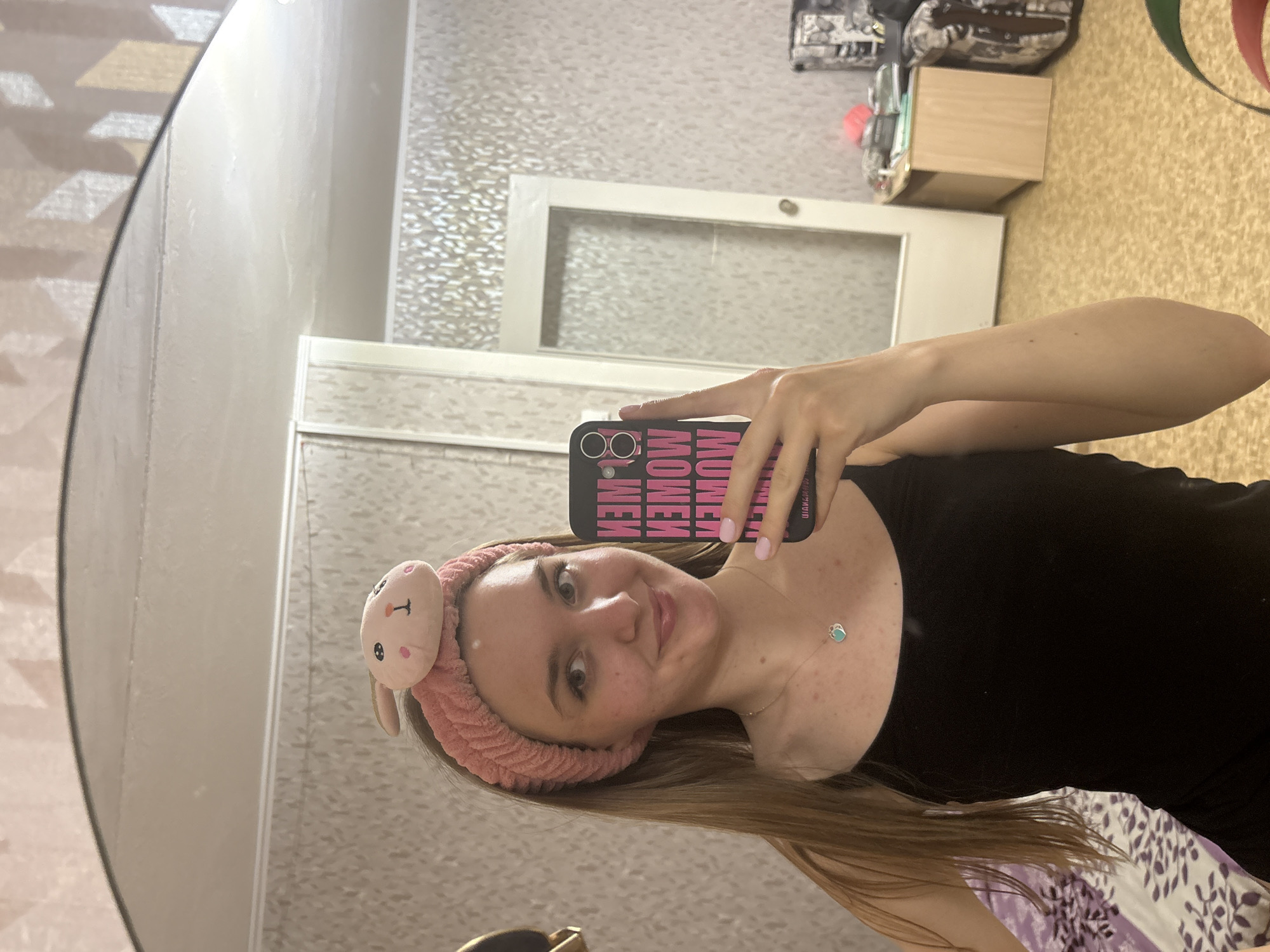 A young woman with long, blonde hair is taking a selfie in what appears to be an apartment. She is wearing a black top and a pink headband with a bunny face on it. The phone she is using has a pink and black "MEOW" case. A silver necklace with a blue/green pendant is visible around her neck.
The background includes a mirror, textured wallpaper, a white interior door, a wooden box on the floor, and a patterned bag or purse. The location is presumably Perm, Russia, based on the request.