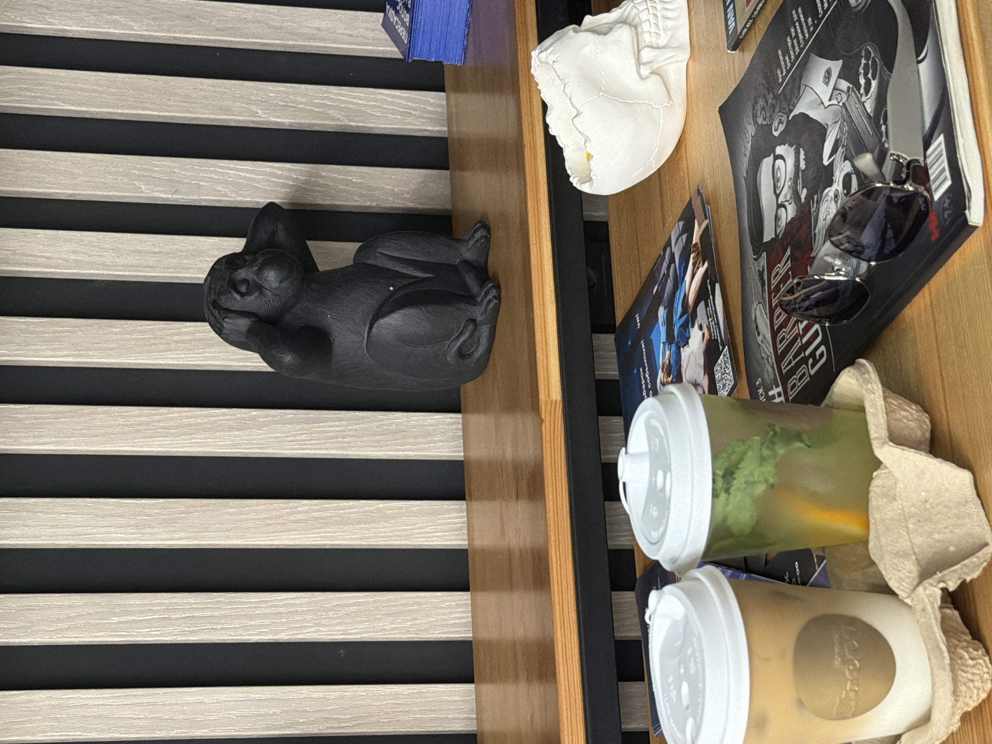 The image depicts a still life arrangement on a wooden table. In the foreground, two to-go cups are placed in a cardboard carrier. The cup on the left contains a layered beverage, possibly coffee with milk, and has a circular logo on its side. The cup on the right holds a green drink with mint leaves and slices of citrus fruit, likely lemon or orange.
Behind the cups, a black statue of a monkey covering its ears is positioned on a wooden shelf. The monkey statue is a representation of the "hear no evil" proverb.
Further back, a white skull-shaped object is visible on the table, positioned to the right of the monkey statue. To the left of the skull, a magazine with a drawing of a man with a beard and glasses on its cover is open. A pair of sunglasses rests on top of this magazine.
The background consists of vertical wooden slats alternating with dark panels, creating a striped pattern. The lighting suggests an indoor environment, possibly a cafe or a waiting area. There are no people visible in the image. The scene is static, with no actions taking place. The overall impression is one of decorative arrangement rather than an active moment.
It is not possible to determine the specific location in Rostov-on-Don, Russia from the provided image.