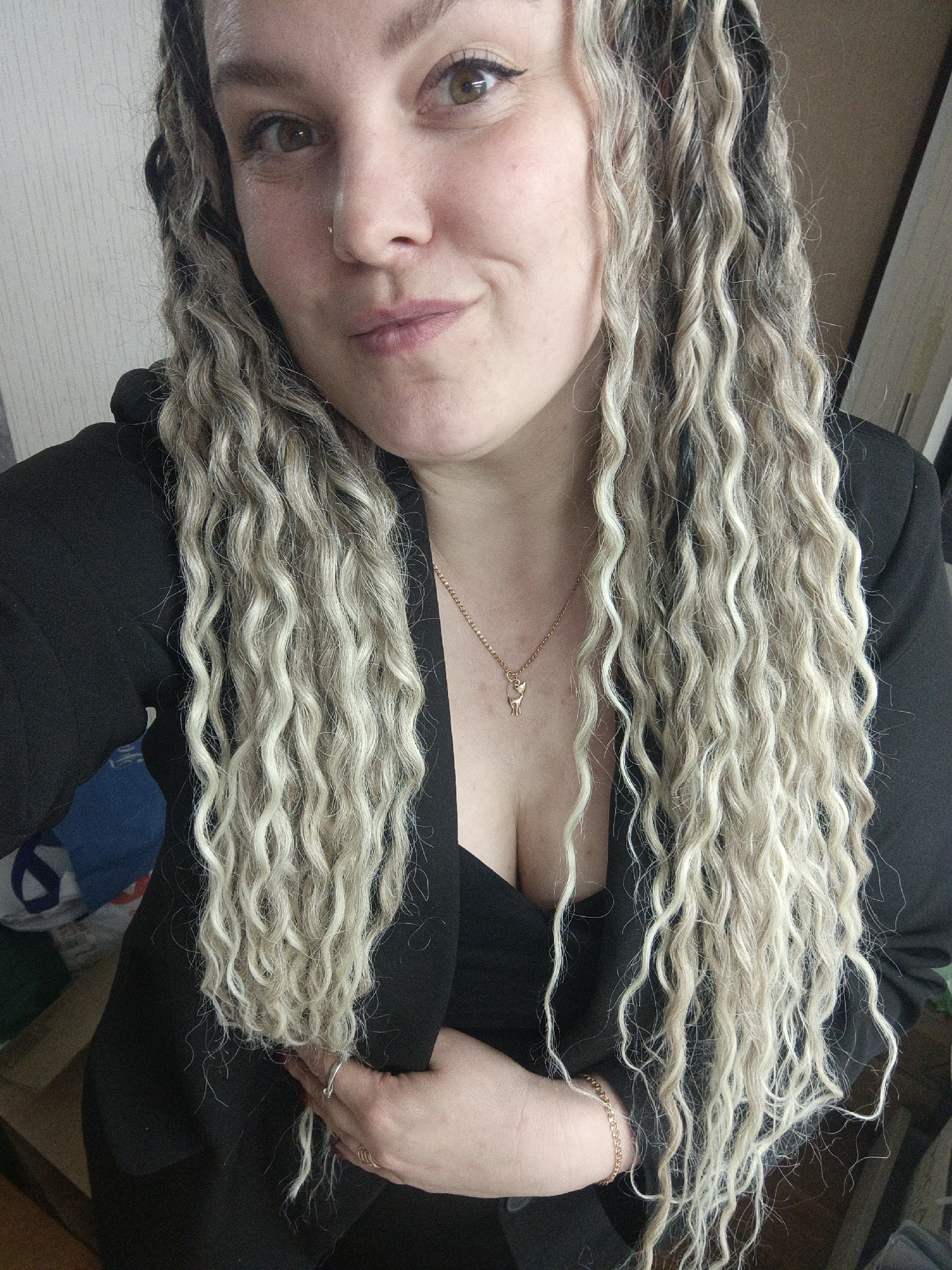 A single adult female is depicted from the chest up, facing the camera with a slight smile. She has long, multi-toned blonde and dark curly hair, likely dreadlocks or similar extensions, cascading over her shoulders. Her eyes are brown, and a small metallic stud is present on her right nostril. She is wearing a black blazer or jacket over a black garment, revealing a décolletage. A gold-colored chain necklace with a small deer-shaped pendant is visible around her neck. Her left hand, with dark-painted fingernails, rests on her chest area; a silver-colored ring is on her ring finger, and a gold-colored chain bracelet is on her wrist. The background is an out-of-focus indoor environment, showing a light-colored, vertically textured wall on the left and a darker, indistinct area on the right, possibly a doorframe. Several blurred objects, including a blue item, are partially visible in the lower-left background. No visual cues within the image indicate the specific city of Noviy, Russia.