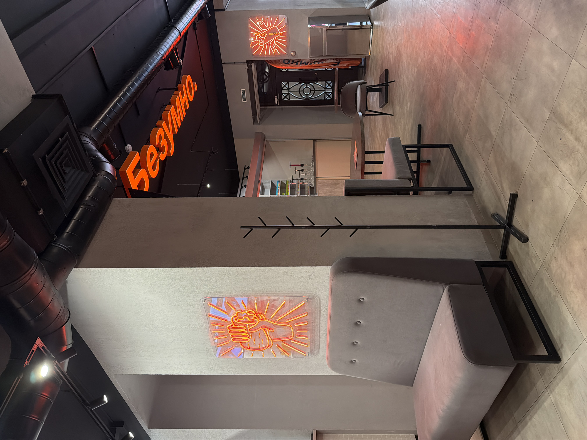 The image depicts the interior of a space in Krasnoyarsk, Russia, specifically inside the "Безумно" restaurant, as evidenced by the prominent neon sign. The restaurant features modern design elements, including exposed ventilation ducts and a neon sign of a fist. The interior includes a seating area with a gray sectional sofa and a metal frame. Additional seating consists of metal-framed chairs and bar stools. The flooring is tiled. A doorway and signage in Russian indicate the exit. Shelves with beverages and glassware are present.