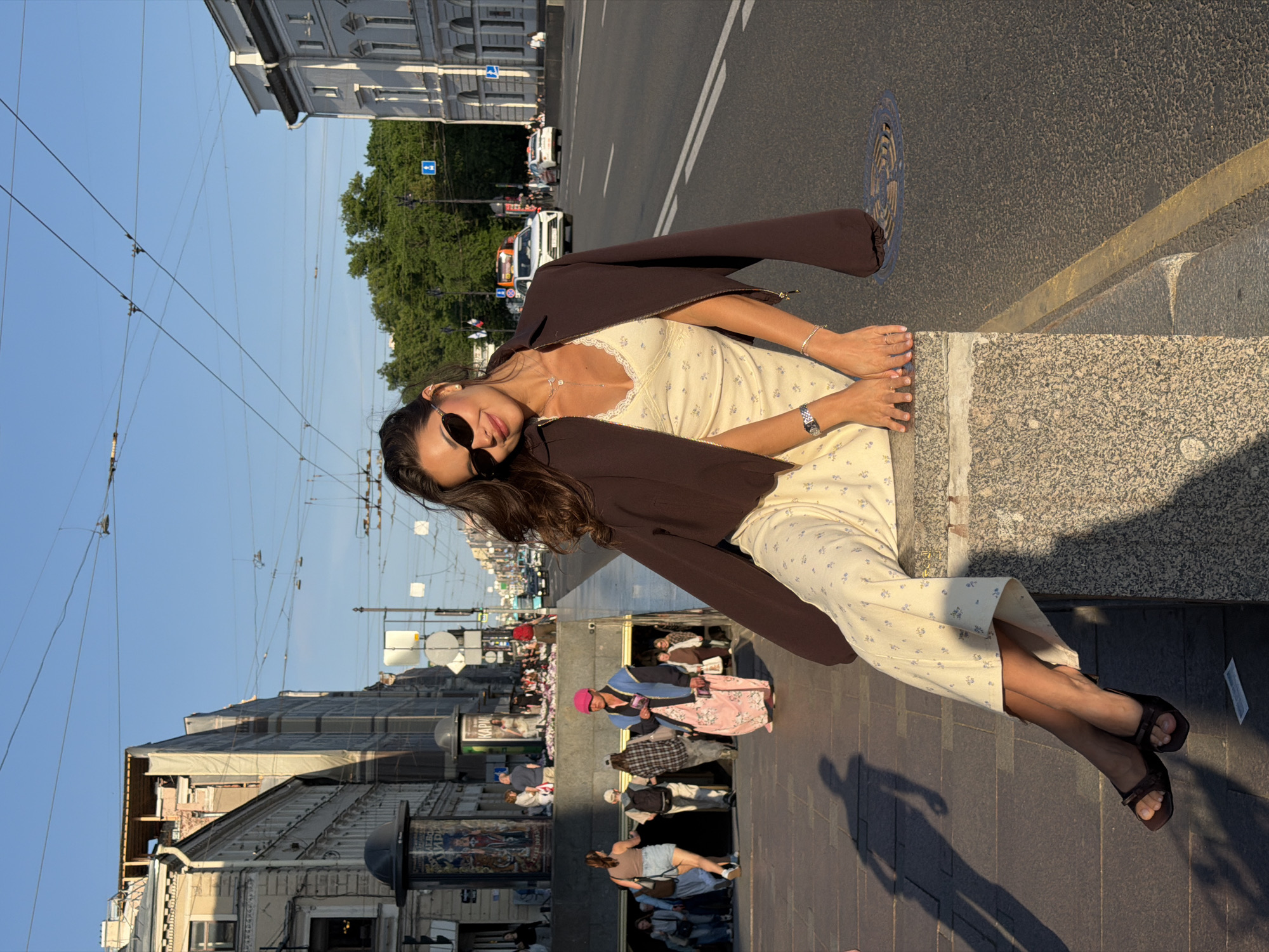 A young woman with long brown hair, wearing dark sunglasses, a light yellow dress with a floral pattern, and a dark brown jacket, is sitting on a stone ledge outdoors in the city of Saint Petersburg, Russia. She is looking towards the camera with a smile. Her hands are resting on her lap, and she is wearing a watch and a bracelet. Her legs are crossed, and she is wearing dark brown sandals.
The background shows a street scene with buildings, cars, and overhead wires for trams or trolleybuses. There are people walking in the background near an entrance to an underground passage. The sun is setting, casting long shadows on the ground, indicating it is late afternoon or early evening. The architecture visible in the background suggests a historic European city. The woman is positioned in the foreground, with the busy city street and pedestrian traffic behind her.