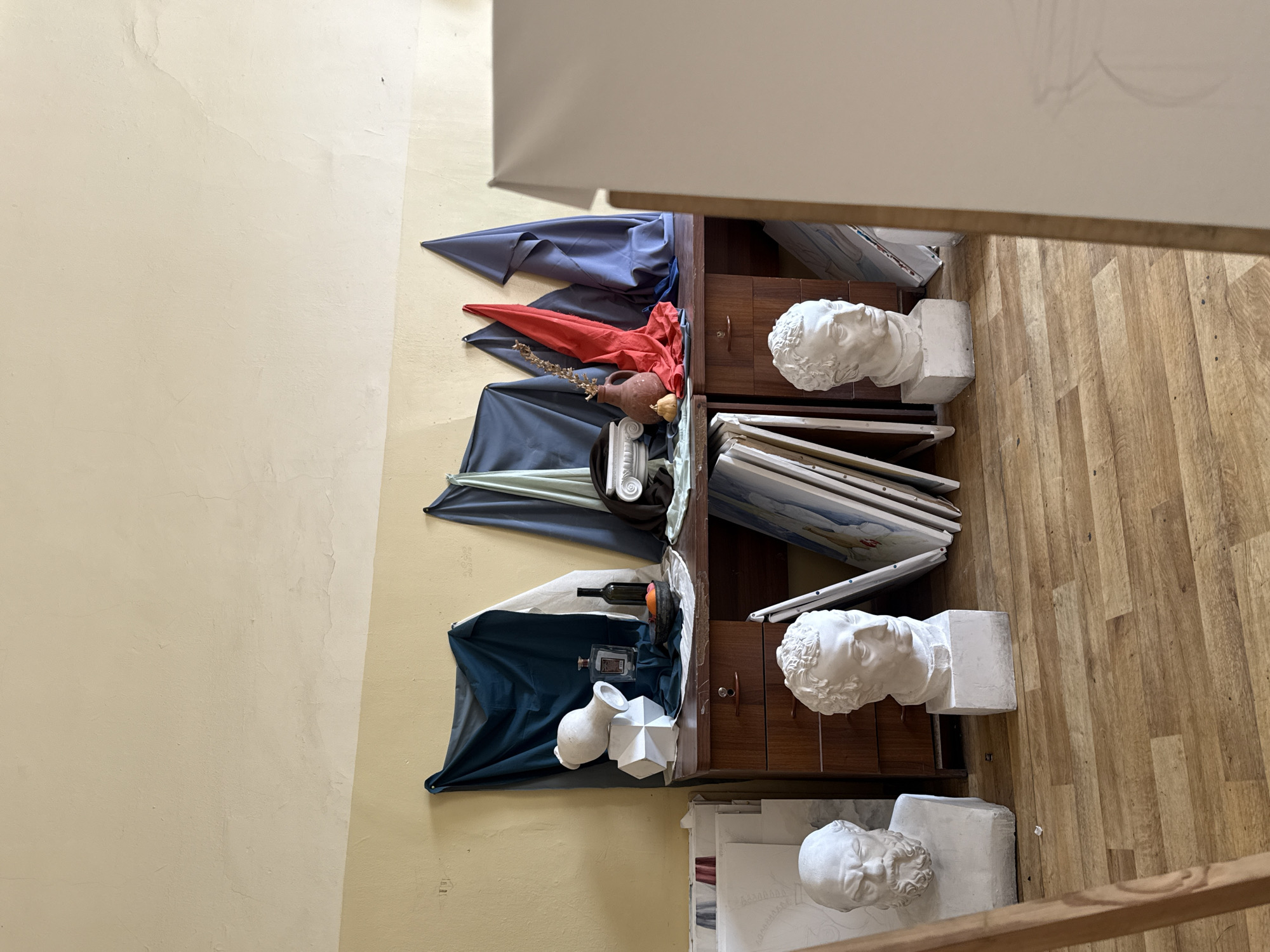 The image depicts a still life arrangement inside a room. The location is presumed to be in Yerevan, Armenia, based on image context.
Arrangement: Three sets of objects are displayed on tiered shelves or cabinets.
Upper Set: A navy blue and red drapery with a brown vase and dried sprigs. There's a portion of what appears to be a classical column also present.
Middle Set: A dark blue drape with a white vase, a polyhedron, a bottle, and a dark object that could be fruit.
Lower Set: A white vase and a polyhedron are placed on a navy drape.
Other Objects: Three plaster head busts of classical figures are visible on the shelves. A stack of paintings leans against the shelf.
Environment: A light yellow wall can be seen behind the furniture. The floor is made of wood planks. A portion of a wooden easel is visible.