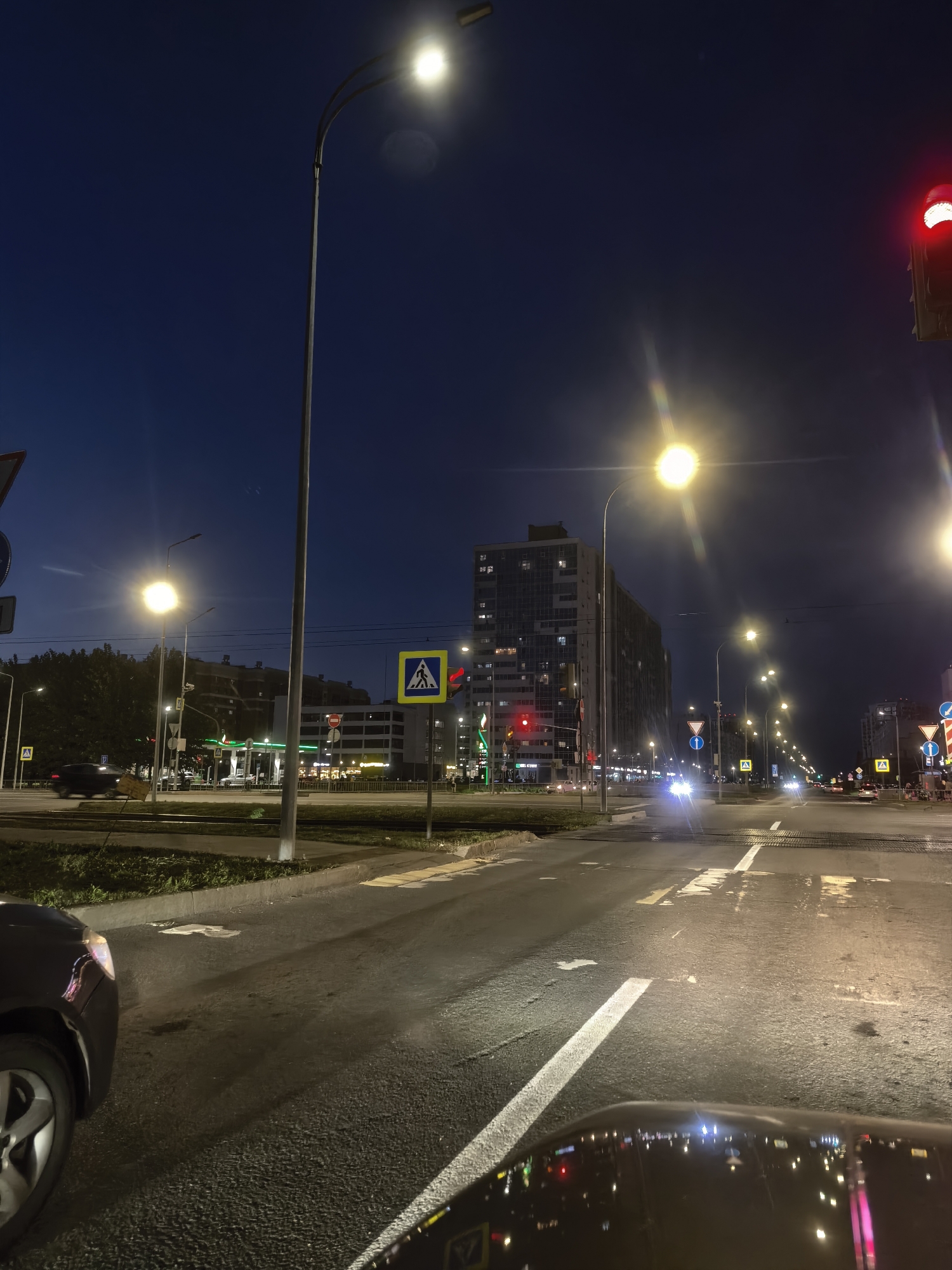 A nighttime, eye-level, wide-angle shot captures a street scene in an urban environment. The sky is a deep twilight blue, transitioning to black at the top. Streetlights illuminate the wet asphalt road, which reflects the lights. In the foreground, the hood and windshield of a dark-colored car are visible. The car is on the right side of the road, facing away from the camera.
The street is lined with trees on the left, behind which are buildings. A tall, modern, multi-story apartment building with numerous windows is prominent in the middle ground. Across the street, a gas station with illuminated pumps and signage is visible on the left. Traffic lights and various road signs, including pedestrian crossing signs and warning signs, are present. Several vehicles are on the road, some with their headlights on, creating streaks of light. The scene depicts a typical urban street at night, with artificial lighting and ongoing vehicle traffic.