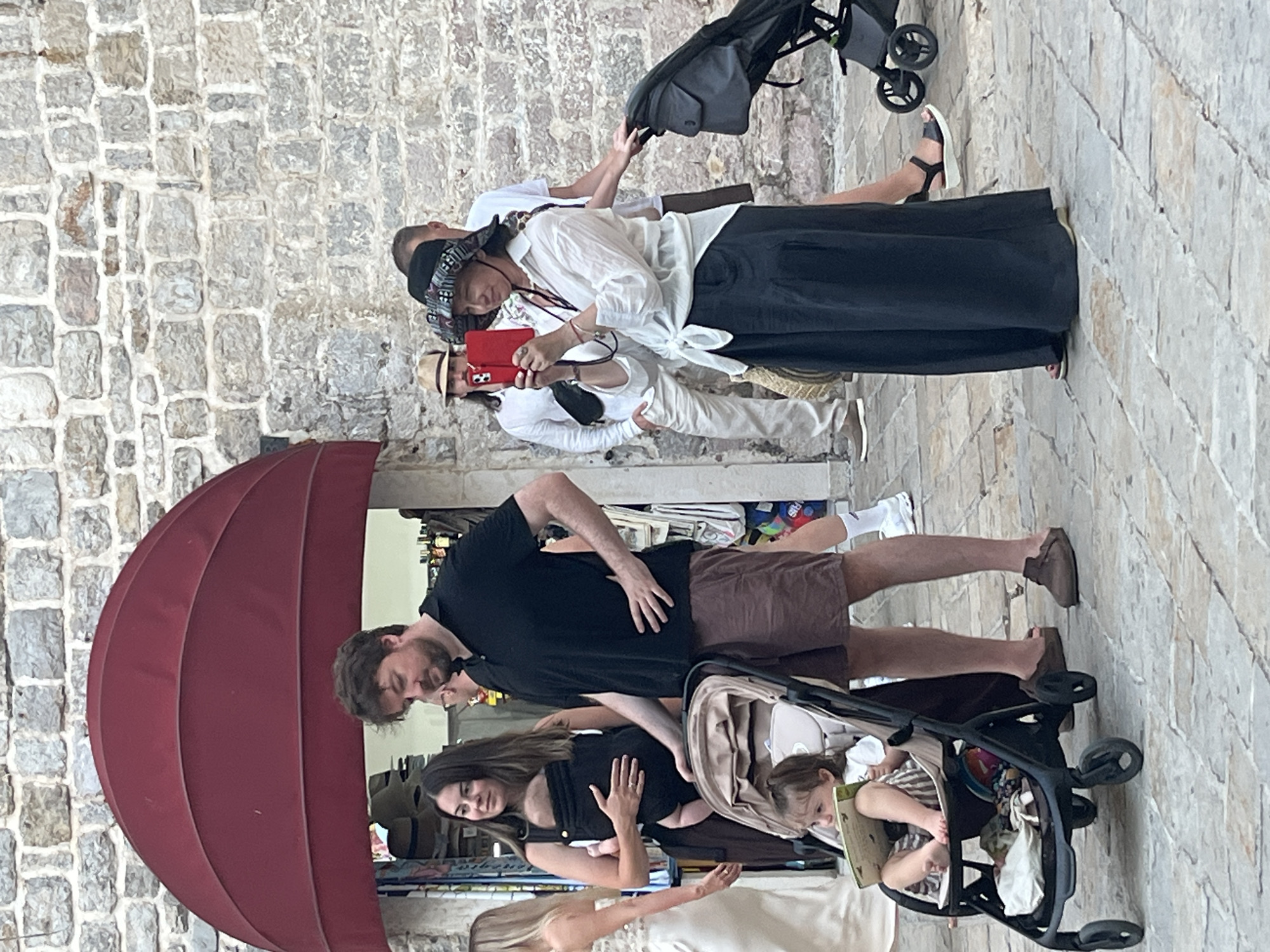 In Budva, Montenegro, a group of people are seen on a cobblestone street in front of a stone building with a large red awning. A man in a black polo shirt and brown shorts stands next to a woman holding a baby in a carrier and next to a stroller. A young child sits in the stroller, reading a book. Another woman in a white shirt and black skirt is taking a photo with her phone, and a man in a white linen outfit is standing behind her. A stroller is also visible to the right. The scene suggests a casual outing on a warm day.