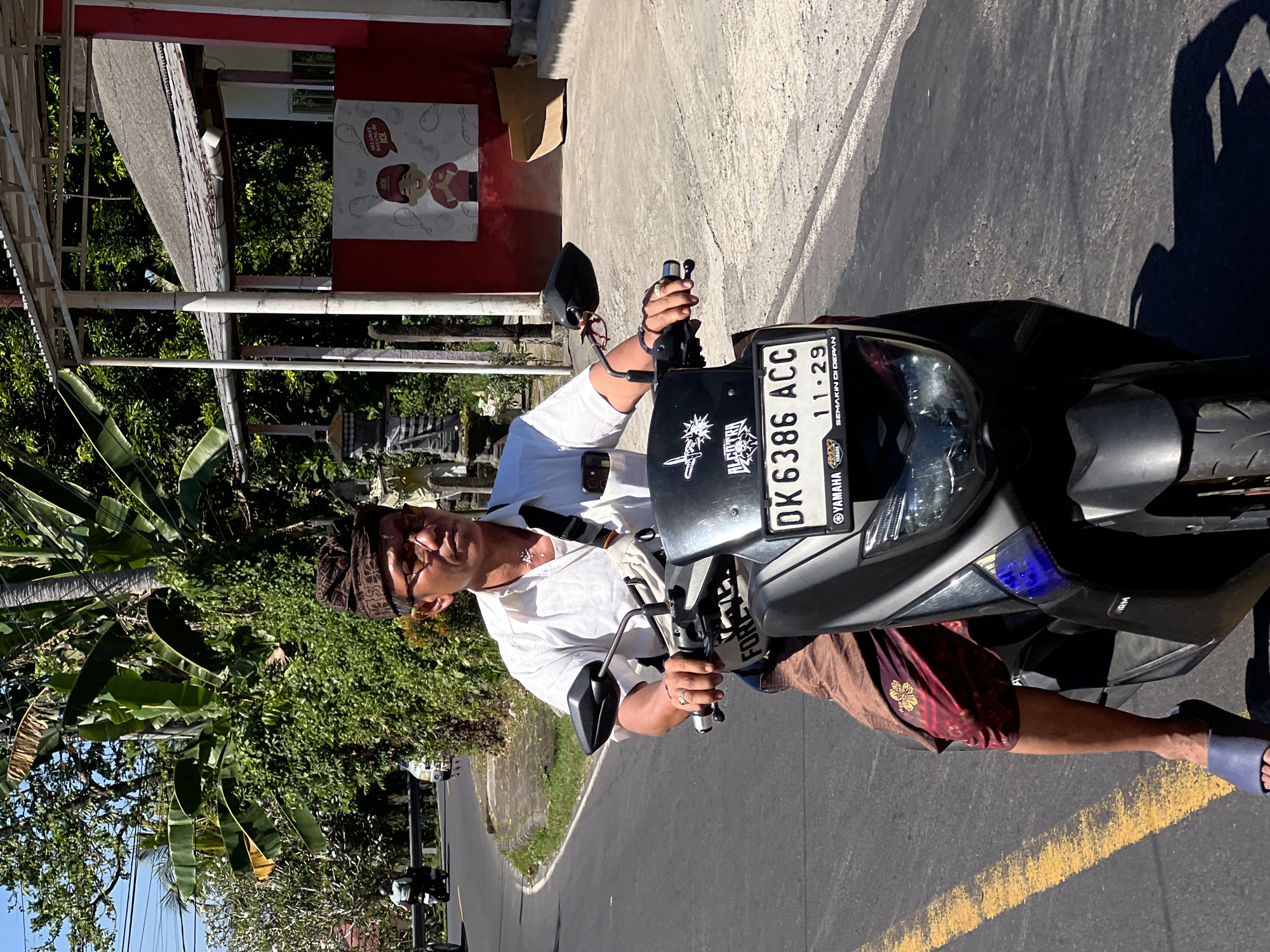 In Manggis, Indonesia, a man with glasses and a patterned head covering is riding a dark gray Yamaha scooter on a paved road under a clear blue sky. He is wearing a white short-sleeved shirt, brown pants, and blue flip-flops. A light-colored bag is slung across his chest. The scooter has a license plate reading "DK 6386 ACC" and various stickers. The road has a yellow line on the left side. Lush green foliage and trees line the right side of the road, along with a building with a red facade and a gray roof. In the background, other vehicles and palm trees are visible. The overall impression is a sunny day scene in a tropical environment.