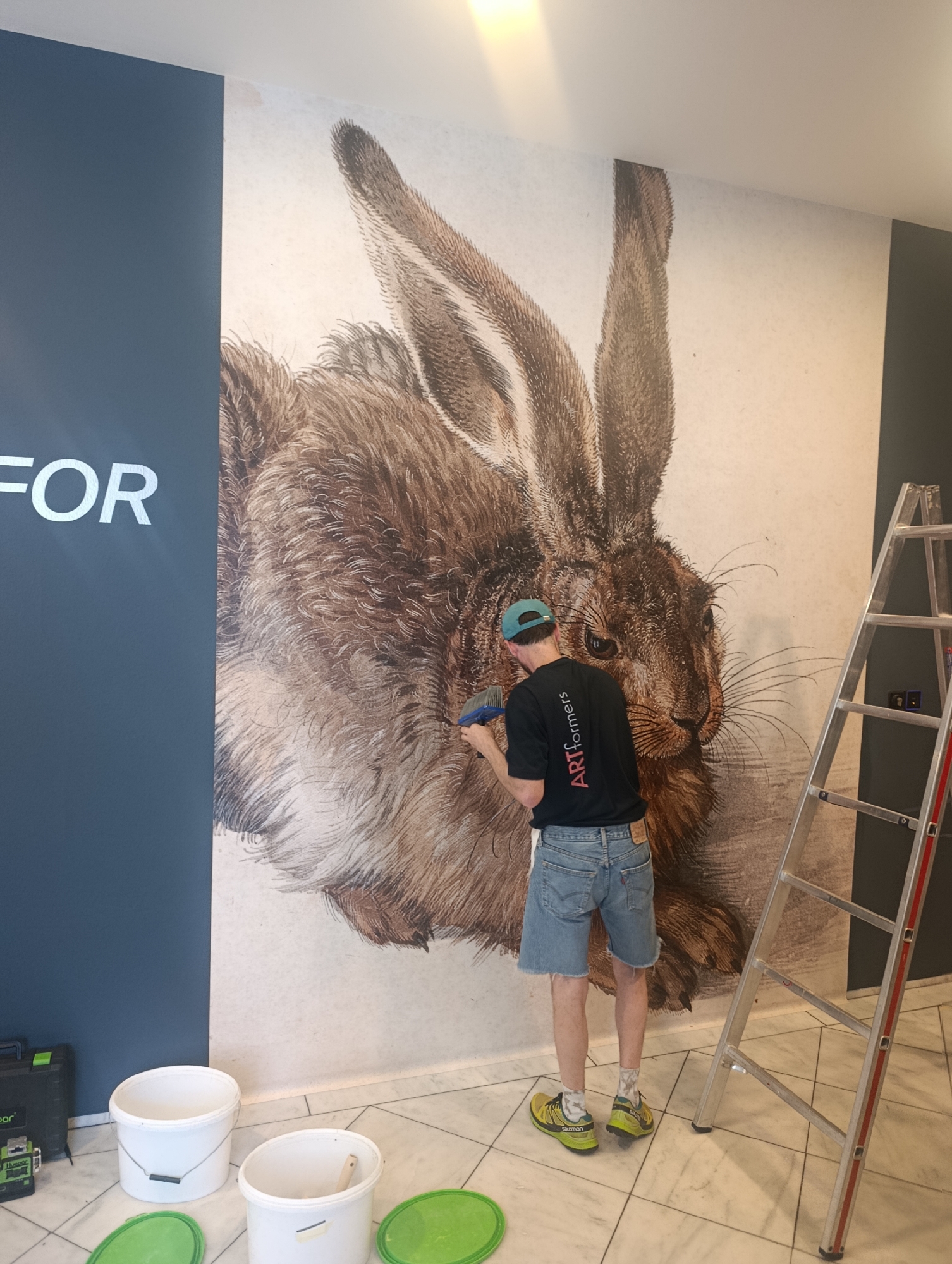 A man is applying a large mural of a hare to a wall. The mural is detailed, with the hare's fur rendered in brown and white. The man is wearing a black t-shirt with "ARTformers" printed on the back, blue denim shorts, a blue baseball cap, and yellow athletic shoes. He is holding a brush and applying the mural to the wall. To the left of the mural, part of a dark blue wall is visible with the white text "FOR" also visible. A silver ladder stands to the right of the mural. In the foreground, there are two white buckets and two green lids on the marble floor, along with a black case with a green light. The scene appears to be indoors, possibly a commercial space, given the scale of the mural and the presence of tools. The lighting suggests it is daytime. The location context is Vienna, Austria.