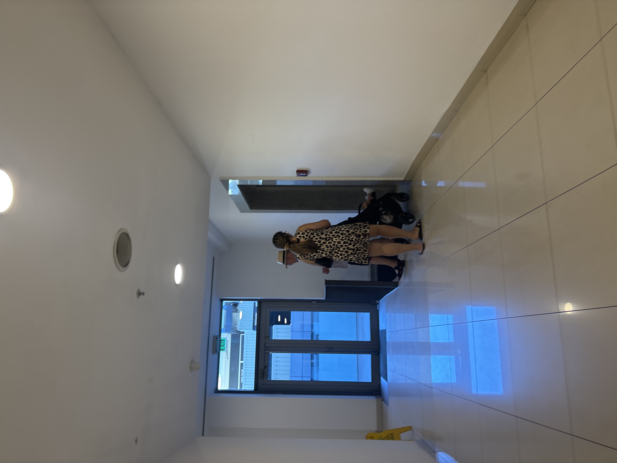 A long, well-lit, tiled hallway leads towards a set of double glass doors. An "EXIT" sign is visible above the doors, which are reflecting a bright blue light from outside, possibly indicating sunlight or artificial lighting on the other side. Two people are standing near the doors; a man wearing a hat and a woman in a leopard-print dress. The woman is pushing a stroller. To the left of the doors, a yellow "wet floor" sign is propped up. The walls of the hallway are white, and recessed lighting illuminates the ceiling. The floor is tiled with light-colored square tiles, with thin dark grout lines. The overall impression is that of a modern, clean, and functional indoor space, likely a hotel or commercial building in Budva, Montenegro.