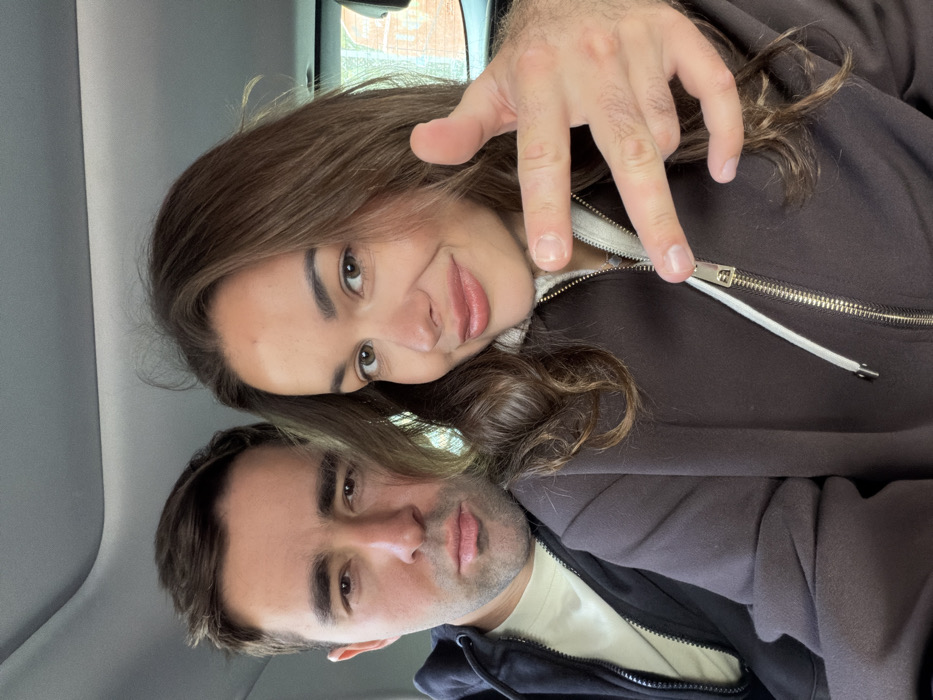 The image displays two individuals, an adult male and an adult female, seated within a vehicle's passenger compartment, identified as being in Tel-Aviv, Israel. The male, positioned on the left, has short dark hair and a neutral facial expression, wearing a dark hooded jacket over a light-colored undershirt. The female, on the right, has wavy brown hair and a slight smile, dressed in a dark brown full-zip hooded sweatshirt with a lighter cream-colored inner lining visible at the collar. The male's right hand is positioned in front of the female, forming a gesture with the thumb and pinky finger extended, while the other fingers are curled. The vehicle's interior headliner is visible above them, and through the rear window, blurred elements of outdoor greenery and reddish-brown building structures are discernible, indicating a daylight, urban or suburban environment.