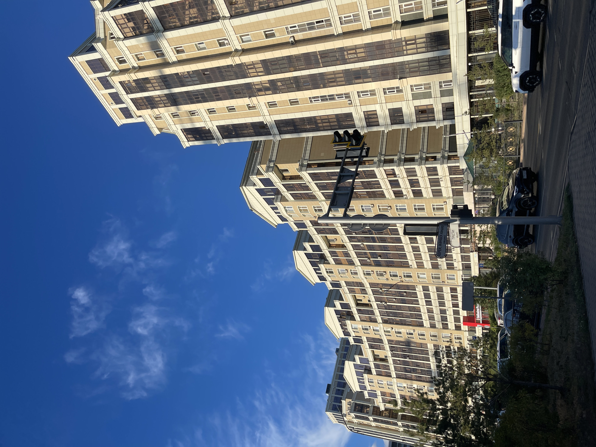 The image shows a street scene in Astana, Kazakhstan, with a row of modern, multi-story apartment buildings dominating the background. The buildings are light-colored with dark-tinted windows and ornate architectural details, particularly at the top. The sky is a clear blue with wispy white clouds. In the foreground, there is a street with several cars, including a black SUV and a white SUV, driving or parked. Trees and bushes line the sidewalk. A traffic light and street signs are visible, one of which has text in what appears to be Cyrillic script. The lighting suggests it is daytime, possibly late afternoon, given the long shadows.