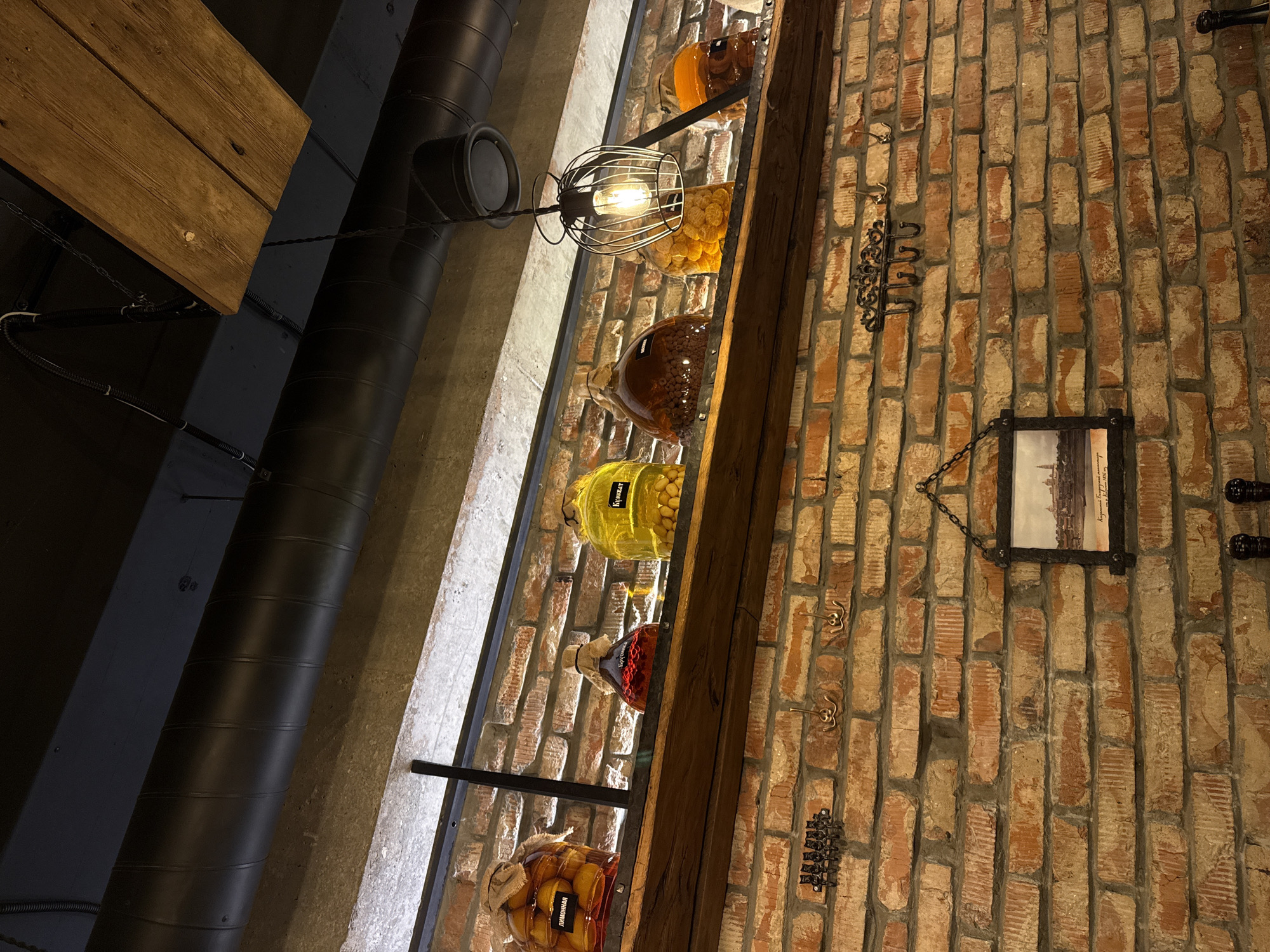 This is a medium-angle, eye-level shot looking up at a rustic interior, likely a cafe or restaurant, with exposed brick walls and wooden accents. The foreground is dominated by a textured red brick wall. Mounted on the wall are several decorative hooks. To the right of the hooks, a framed black-and-white photograph hangs, depicting a city skyline with prominent domed buildings, possibly of Kazan, Russia, as indicated by the Cyrillic inscription below.
Above the brick wall, a wooden shelf runs horizontally, displaying a collection of large glass jars filled with various preserved items. From left to right, the first jar contains what appear to be whole yellow fruits, possibly lemons, submerged in liquid, with a label reading "Лимонная" (Lemonnaya). The second jar is reddish-brown and appears to contain red berries, possibly cranberries or a similar fruit. The third jar is yellow and filled with small, round, yellow objects, labeled "Кульбаб" (Kul'bab), which translates to dandelion. The fourth jar is a dark amber color, filled with small, dark, round items, possibly nuts or seeds. The fifth jar contains bright yellow, circular objects, possibly dried apricots or similar fruit. The sixth jar, partially obscured on the far right, contains amber-colored preserved items.
Above the shelf, a black metal duct pipe curves across the frame, supported by visible industrial elements. A wooden beam is also visible near the top of the image, extending from the ceiling. A decorative light fixture with a cage-like shade hangs from a wire, illuminating the scene with a warm glow. The overall impression is one of rustic charm with a focus on preserved food items.