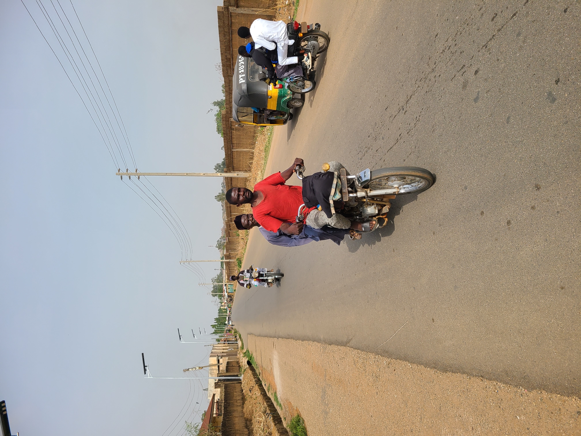 This daytime image captures a scene of everyday transportation on an asphalt road in Barakin Lerawi, Nigeria. The sky is bright and clear, suggesting sunny weather.
In the foreground, a man wearing a red shirt and a dark cap is driving a silver-colored motorcycle, with another person, partially obscured, riding behind him in a light blue garment. They appear to be moving towards the left side of the frame.
Further down the road, another motorcycle carries multiple passengers, including one person in a white garment and another in a dark blue top. Alongside this motorcycle is a "Keke NAPEP" (auto-rickshaw) in green, yellow, and black, displaying the text "PT-18XG" on its side. In the distance, more motorcycles and people are visible, indicating traffic flow.
The road is bordered by dusty shoulders and low-rise buildings, some featuring bare brick construction. Utility poles with power lines run parallel to the road on both sides, extending into the background. Modern streetlights are also present along the roadside. The scene depicts a typical busy thoroughfare with people commuting on two-wheeled and three-wheeled vehicles.