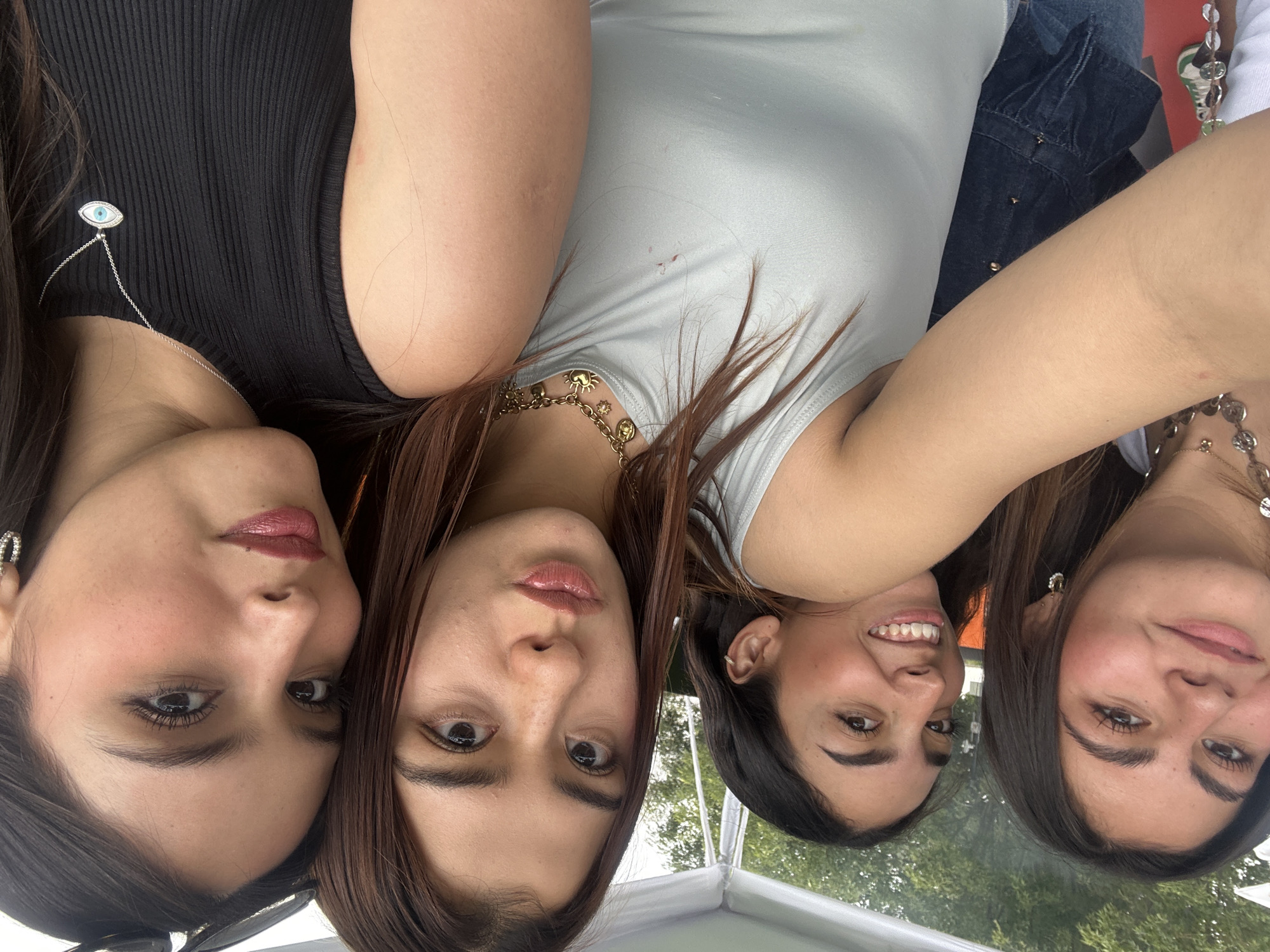 Four women are huddled together, likely taking a selfie. The woman on the left wears a black top and a necklace with an eye pendant. The next woman wears a gold necklace. The next woman is smiling. The woman on the right wears a necklace with round pendants. The image orientation is inverted. The background shows glimpses of green foliage, possibly trees. The location is potentially San Bernardino Tlaxcalancingo, Mexico.