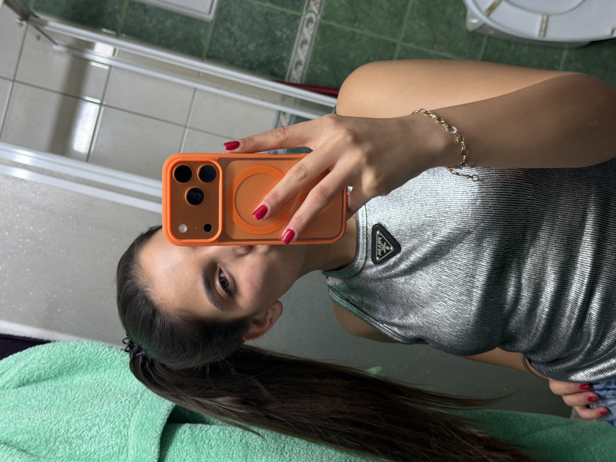 A young woman with long dark hair pulled back in a ponytail is taking a selfie in what appears to be a bathroom. She is holding an orange iPhone with an orange MagSafe charger attached, covering her face partially. Her nails are painted red. She is wearing a silver, form-fitting top with a Prada logo on the shoulder. She is wearing a gold bracelet on her right wrist. She appears to be standing in front of a light green towel. The bathroom has light green patterned tiles and a white shower/tub. Based on the tiles in the background, it is likely in Rudartsi, Bulgaria.