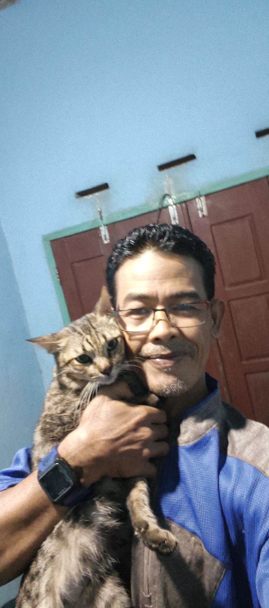 In Muaro Jambi, Indonesia, a man with dark hair, glasses, and a mustache holds a tabby cat. He is wearing a blue and gray jacket, and a dark smartwatch on his left wrist. The cat is held close to his chest, with its head resting on his shoulder. The background shows a light blue wall with two dark horizontal accents, and a dark red door with a green frame. The lighting is dim, suggesting an indoor setting.