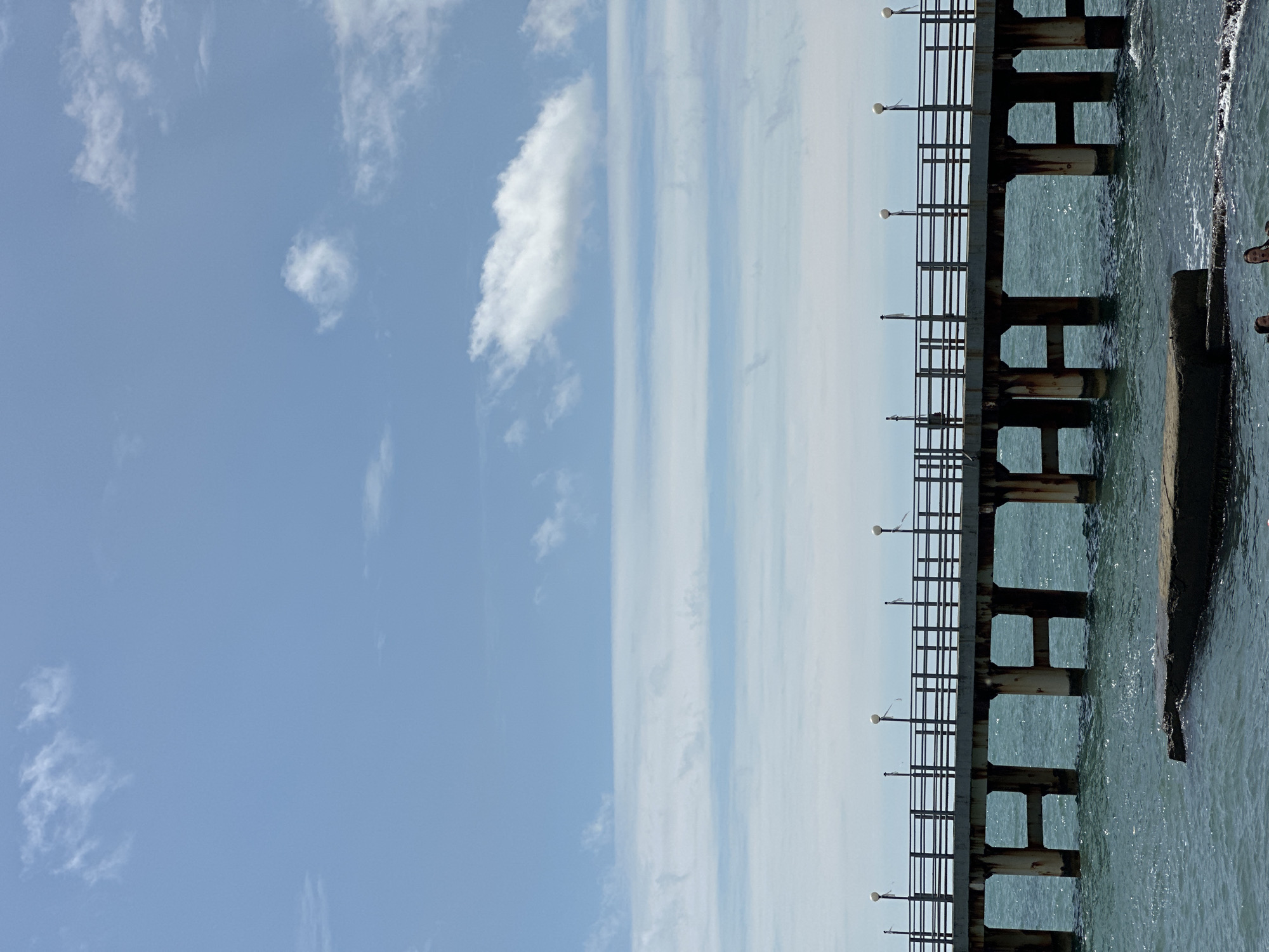 The image depicts a wooden pier extending over a body of water. The pier has a railing along its length and is supported by several concrete pillars. There are lights installed along the railing. The water beneath the pier appears to be choppy. In the foreground, partially submerged concrete structures are visible. The sky above is blue with scattered white clouds, some of which appear to be thin and elongated. The overall scene is one of an outdoor coastal environment. Based on the provided context, this pier is located in Sochi, Russia.
