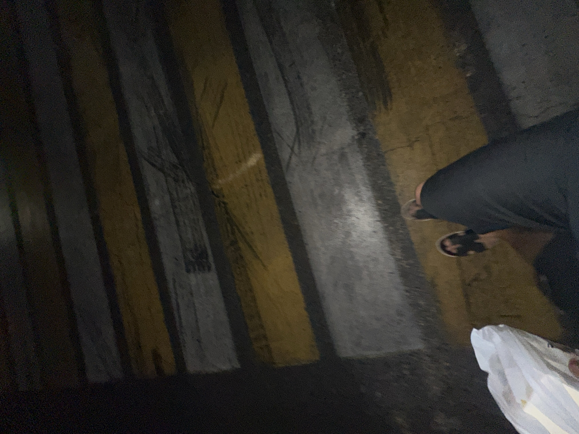 The image shows a person's legs and feet wearing dark pants and sandals, standing on a pedestrian crossing. The pedestrian crossing has alternating yellow and white stripes. The scene appears to be outdoors at night, with artificial lighting casting shadows. The ground is wet, suggesting it may have rained or been recently washed. A white plastic bag is visible on the ground near the person's feet. The overall impression is of a dimly lit urban environment, possibly a street in Tashkent, Uzbekistan, due to the provided location context. The image is taken from a low angle, looking down at the person's feet and the pedestrian crossing. There is no visible text in the image.