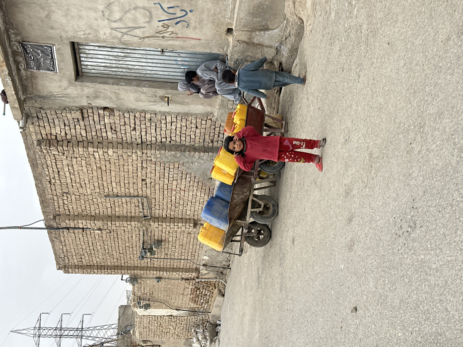 The image portrays an outdoor street scene in Quetta City Tehsil, Pakistan, under an overcast sky. In the immediate foreground, a young child, estimated to be between three and five years old, stands centrally on a light grey paved surface. The child is dressed in a red long-sleeved shirt and red patterned trousers, wearing red footwear, with hands raised to the sides of their head.
Behind the child and slightly to the right, a multi-wheeled cart, resembling a handcart or small trailer, is stationary. It is loaded with at least eight plastic jerry cans: approximately five yellow and three blue.
To the right of the frame, an unpaved section of ground meets a concrete building with a light grey, stucco-like finish. Three individuals are seated on a raised concrete ledge adjacent to this building. An adult, mostly obscured, is present alongside two younger individuals. One child or youth, dressed in a light-colored traditional garment, is seated facing the adult. A second child, wearing a blue outfit, is seen walking away from this group toward the right edge of the frame. The building features a corrugated metal door and a black sign with white script positioned above the door. Graffiti, including the letters "AS" and other script, is visible on the wall to the right of the door.
To the left of the scene, a tall brick wall with exposed utility meters and pipes extends along the street. In the distant background, a prominent metal lattice electricity pylon is visible against the muted sky, accompanied by overhead power lines. The ground surface is uneven and appears to be asphalt or concrete.
