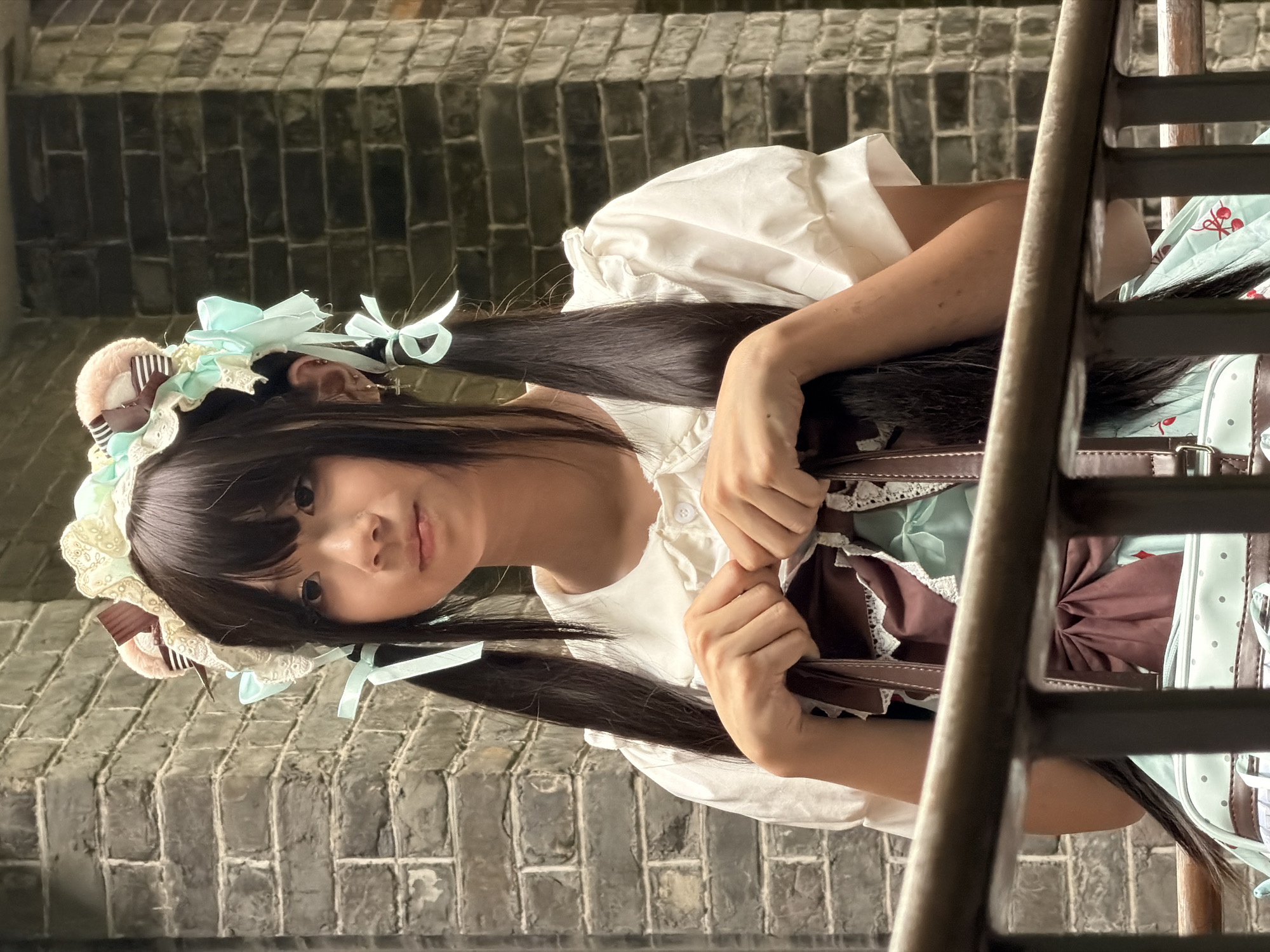 A young woman with long, dark hair styled in pigtails and wearing a lolita-inspired headband with bear ears and lace trim, a white puff-sleeve blouse, and a light blue dress with cherry patterns, is pictured from the chest up, holding a brown handbag with lace trim. She is positioned in front of a dark brick wall and a metal railing. The scene is likely outdoors, possibly in a courtyard or walkway, given the brickwork and railing, suggesting an urban environment within Chancheng District, China, consistent with traditional architecture often found in such areas.