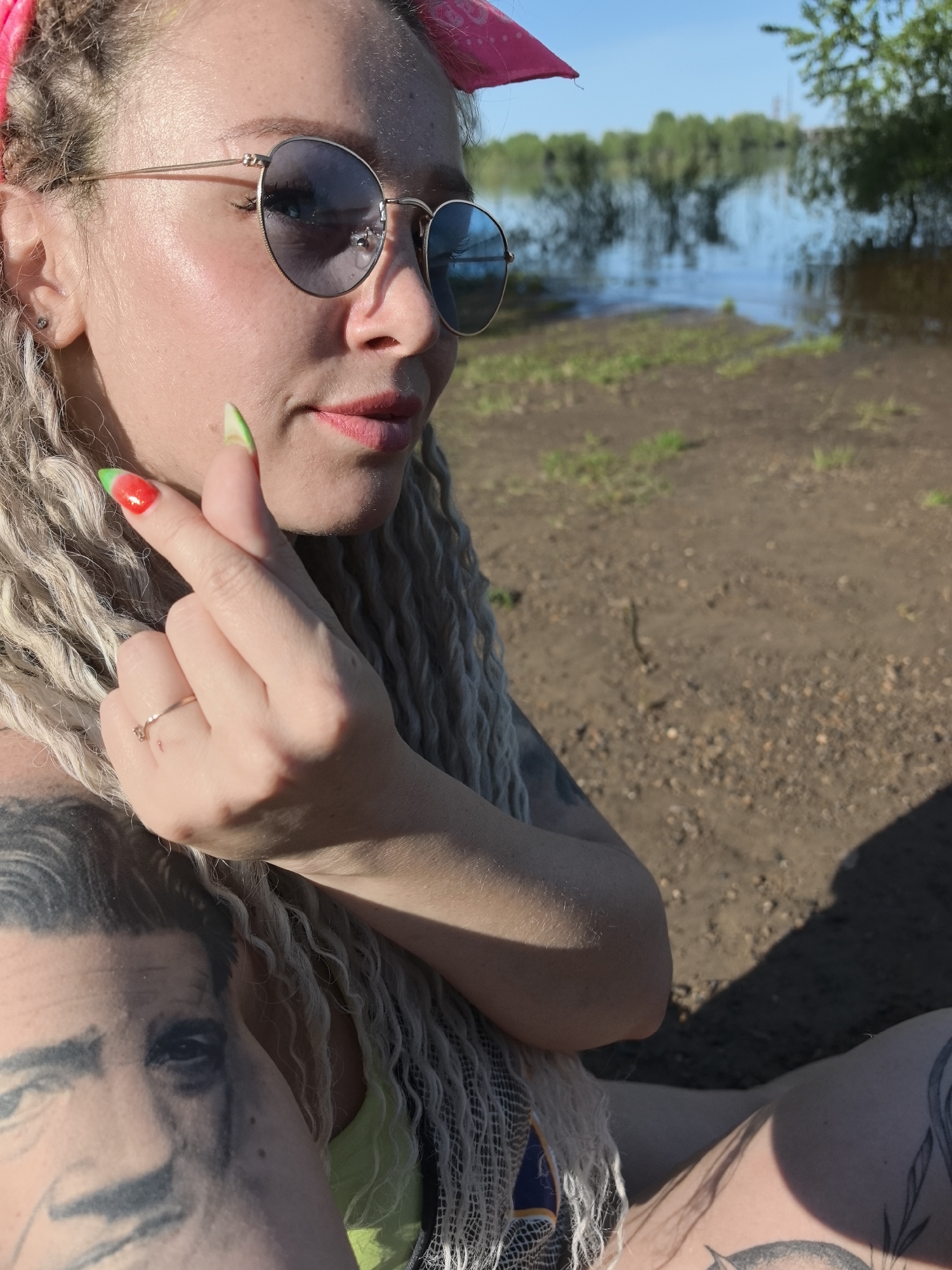A woman with blonde dreadlocks and sunglasses is sitting near a body of water in Noviy, Russia. She has a pink bandana in her hair and multiple tattoos are visible on her arms and legs, including a face tattoo on her upper arm. Her fingernails are painted red and green. The ground appears to be dirt and grass. The body of water is in the background with trees visible on the opposite shore.