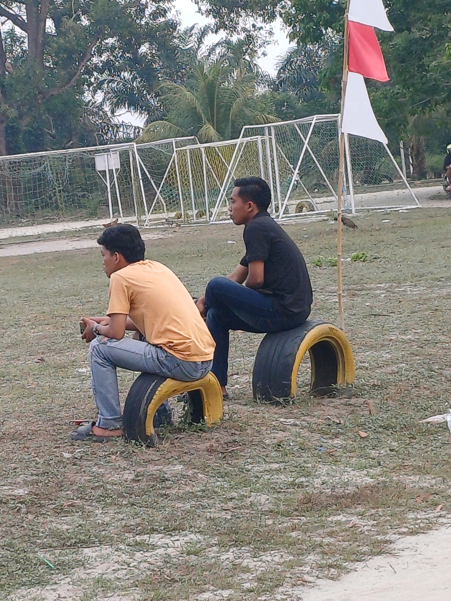 Two young men are sitting on repurposed tires, which serve as seats, in an outdoor grassy area. The man on the left is wearing an orange t-shirt and blue jeans, and is holding a dark object in his hands. The man on the right is wearing a black t-shirt and blue jeans. Behind them, there is a soccer goal with a white net, and a flagpole with two flags, one red and one white. Trees and other greenery are visible in the background. The ground is covered in a mix of grass and dirt. The scene appears to be located in Dumai City, Indonesia, likely in a park or recreational area.