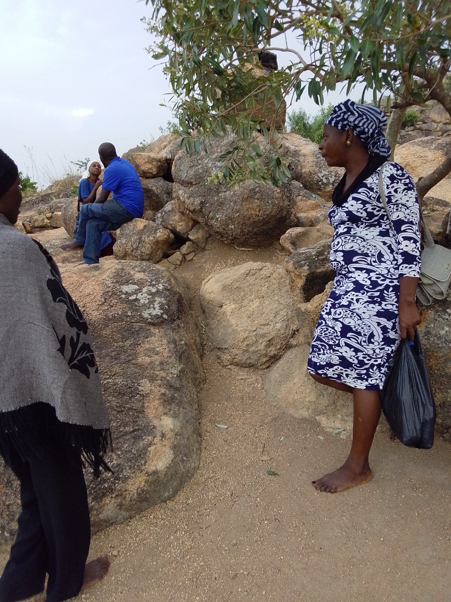 The image shows a group of people in Gura Top, Nigeria, likely outdoors in a rocky area. A woman in a patterned blue and white dress stands barefoot, holding a black plastic bag. She wears a headwrap. Another person is partially visible on the left, wearing a grey shawl and black pants, also barefoot. In the background, two people sit on rocks. One is wearing a blue shirt and jeans. The environment consists of large rocks, sparse vegetation, and a sandy ground surface. A small tree with sparse foliage is visible in the background. The sky is overcast.