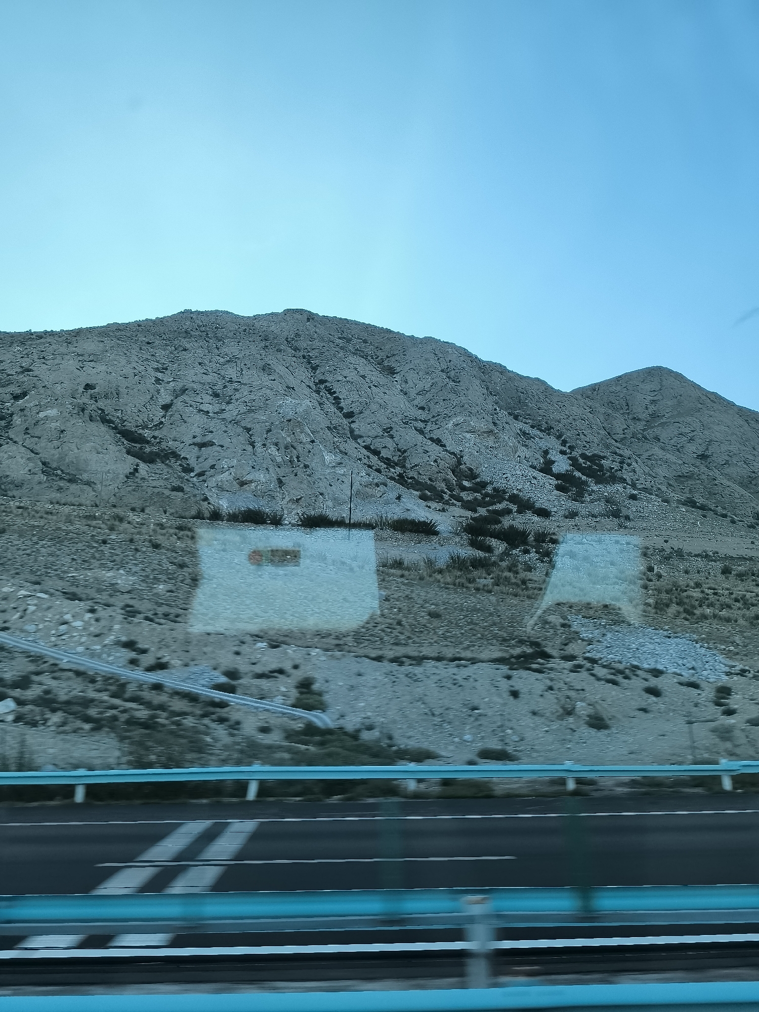 The image shows a mountainous desert landscape seen from a moving vehicle. The foreground features a highway with a blue railing. The mountains in the background are rocky and arid, with sparse vegetation. There are reflections on the glass, indicating the photo was taken through a window. The sky is a clear, light blue. The specific location in China is unknown.
