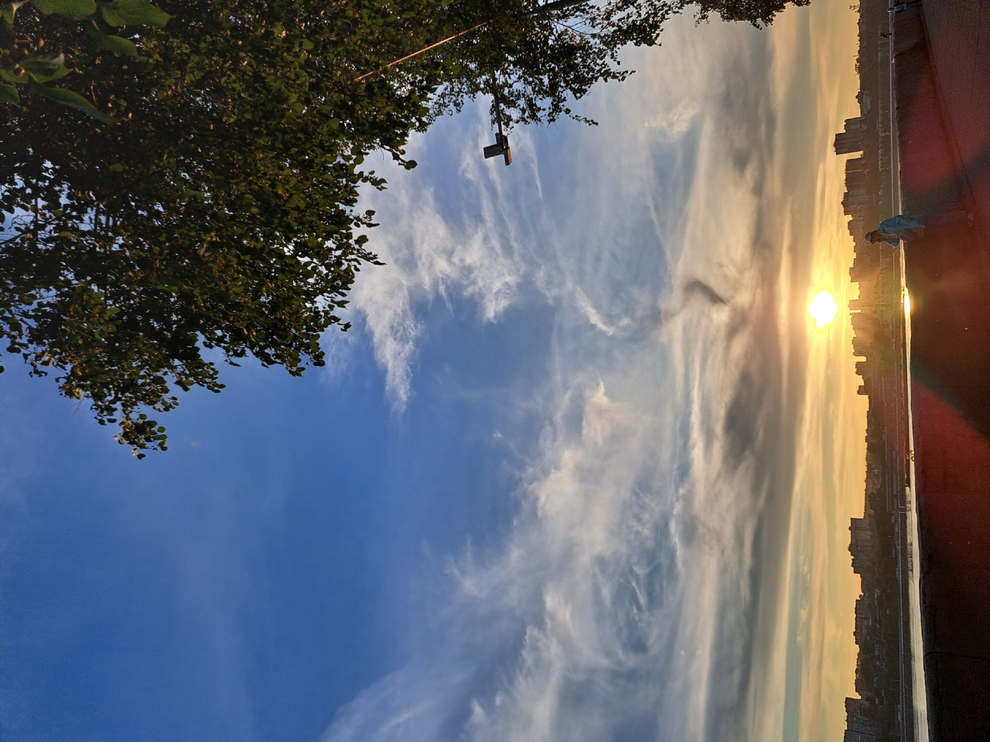 A picturesque sunset casts a warm glow over the cityscape of Irkutsk, Russia, as seen from the embankment of the Angara River. The sky is a vibrant canvas of blue, transitioning to soft oranges and yellows near the horizon, with wispy clouds creating a dynamic celestial display. Silhouetted against the setting sun are numerous buildings that form the Irkutsk skyline. A lone figure, dressed in a light blue jacket and a cap, leans against a dark-colored wall, seemingly engrossed in conversation on their phone. Another person is visible in the distance, further down the embankment. A lush green tree frames the top right corner of the image, its leaves partially obscuring the sky and adding a touch of nature's beauty to the urban panorama. A lamppost is suspended in the sky, positioned to the right of the frame.