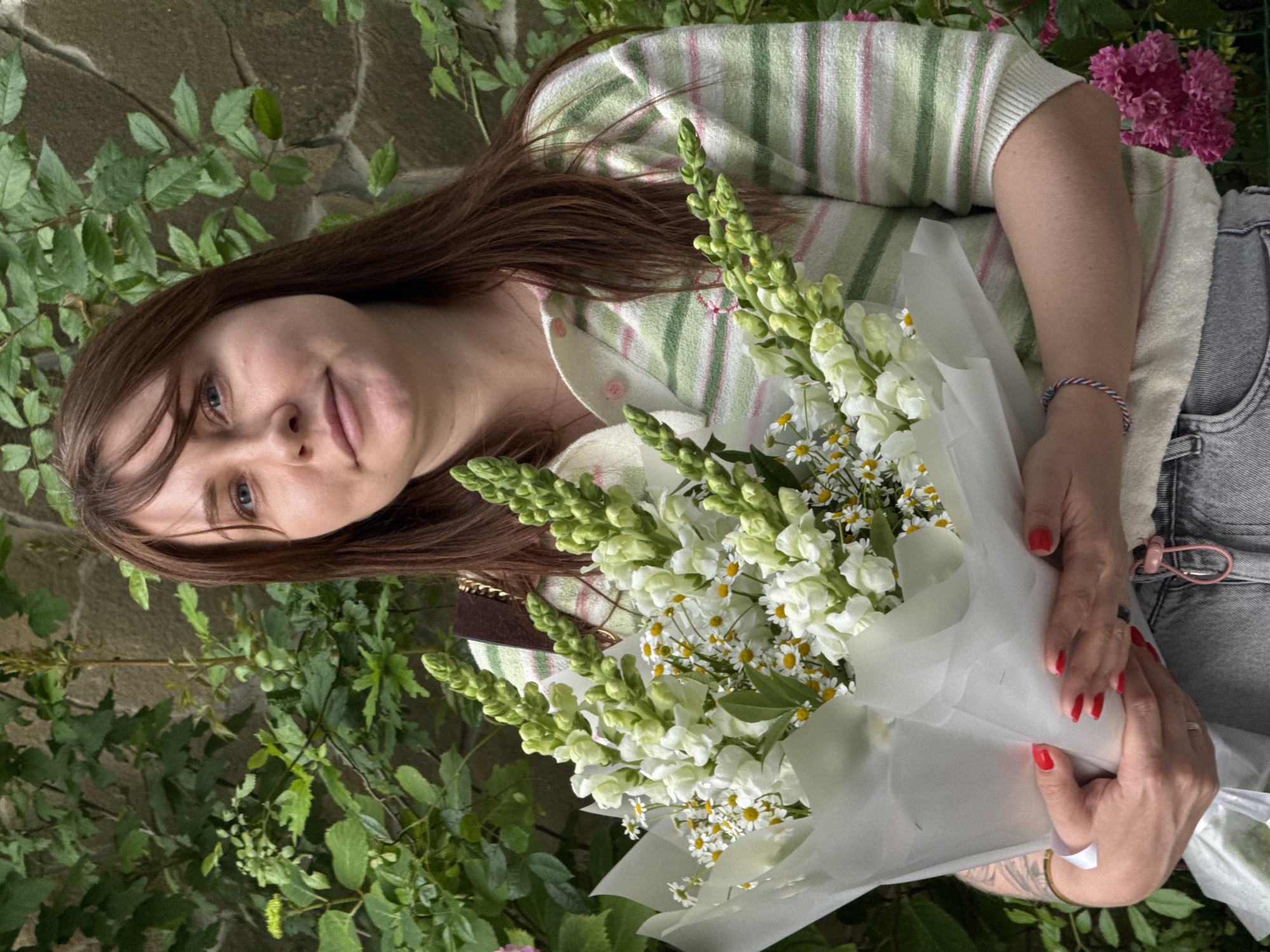 A medium shot captures a young Caucasian woman holding a bouquet of white snapdragons and daisies. She is positioned outdoors against a backdrop of lush green foliage and a stone wall. The woman has fair skin, light blue eyes, and long, dark brown hair with bangs. She is wearing a light-colored, striped knit sweater with green and pink stripes, and gray denim jeans. Her hands, adorned with bright red nail polish, are clasped around the bouquet. The bouquet itself is wrapped in white paper. The scene appears to be in a natural, outdoor setting, possibly a garden, with no specific urban landmarks visible to identify it as Sochi, Russia.