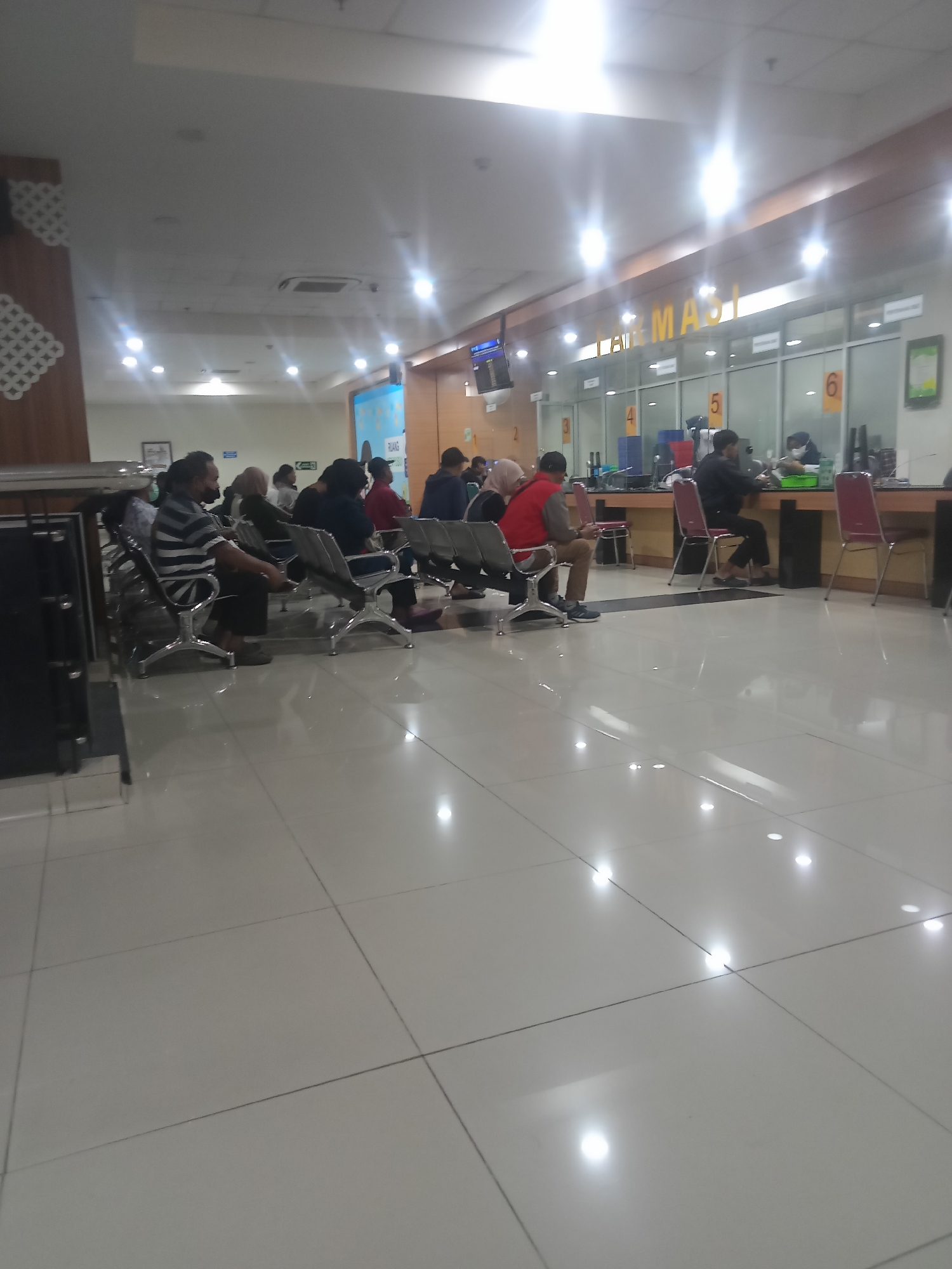 The image depicts the interior of what appears to be a government office or a public service center in Depok, Indonesia. The scene is brightly lit, with a polished tiled floor reflecting the overhead lights. Several individuals are seated in a waiting area, facing a counter with multiple service windows. The counter has large glass partitions, and numerical labels (e.g., 2, 3, 4, 5, 6) are visible above some of the windows, indicating different service points. In the background, a sign with the word "INFORMASI" (Information) is visible. People are dressed casually, and some appear to be engaged in conversations while waiting. The overall atmosphere suggests a routine day at a public service facility.