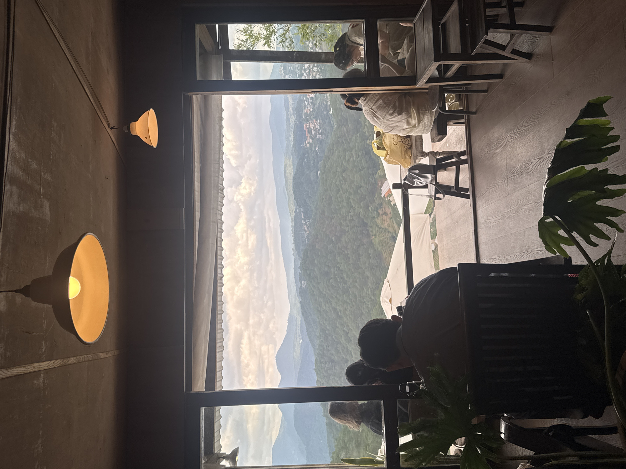 This image depicts an indoor seating area with a panoramic view of a lush, mountainous landscape, characteristic of Xuân Thành, Vietnam. The scene is framed by a large open doorway leading to an outdoor patio. Inside, wooden flooring and dark wooden furniture, including chairs and tables, create a rustic ambiance. Two industrial-style pendant lamps hang from the ceiling, casting a warm glow.
Several individuals are visible. In the foreground, a person sits with their back to the camera, facing a dark wooden chair. To the left, a person with blonde hair is seen from behind, interacting with another individual. Further into the seating area, two people are seated at a table; one person, wearing a light-colored shirt, is facing away from the camera, while the other, a woman with dark hair pulled back, is looking down at something on the table.
The view through the doorway reveals a breathtaking vista of rolling green hills covered in dense forest, extending into the distance. Layers of mountains fade into a hazy sky, suggesting a significant elevation. The sky is partly cloudy with soft, diffused light. On the outdoor patio, several white umbrellas are set up, and the wooden floor continues from the indoor area. A yellow tote bag and a black purse are placed on a stool. The overall impression is one of a serene cafe or restaurant offering a spectacular natural backdrop.