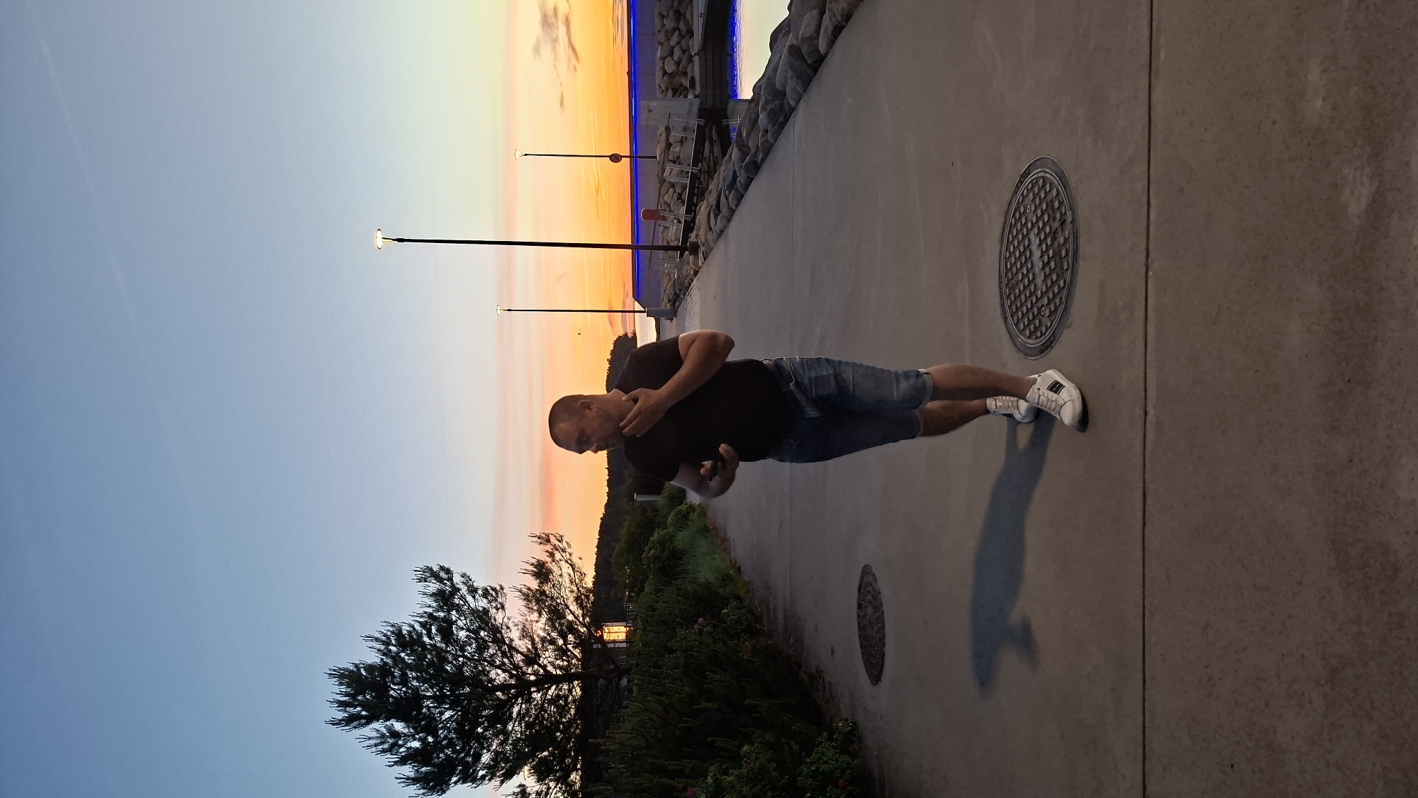 A man is walking on a paved walkway with a view of a harbor during sunset. The man is wearing a black t-shirt and denim shorts, with white sneakers. He is looking to his left, with his hand touching his chin. The walkway is bordered by rocks and a body of water on the right, and a line of trees and bushes on the left. Several streetlights are visible along the walkway. The sky displays a gradient of orange, yellow, and blue, indicating the sunset. The scene appears to be a public area, possibly a promenade or park in Tallinn, Estonia, near the harbor.