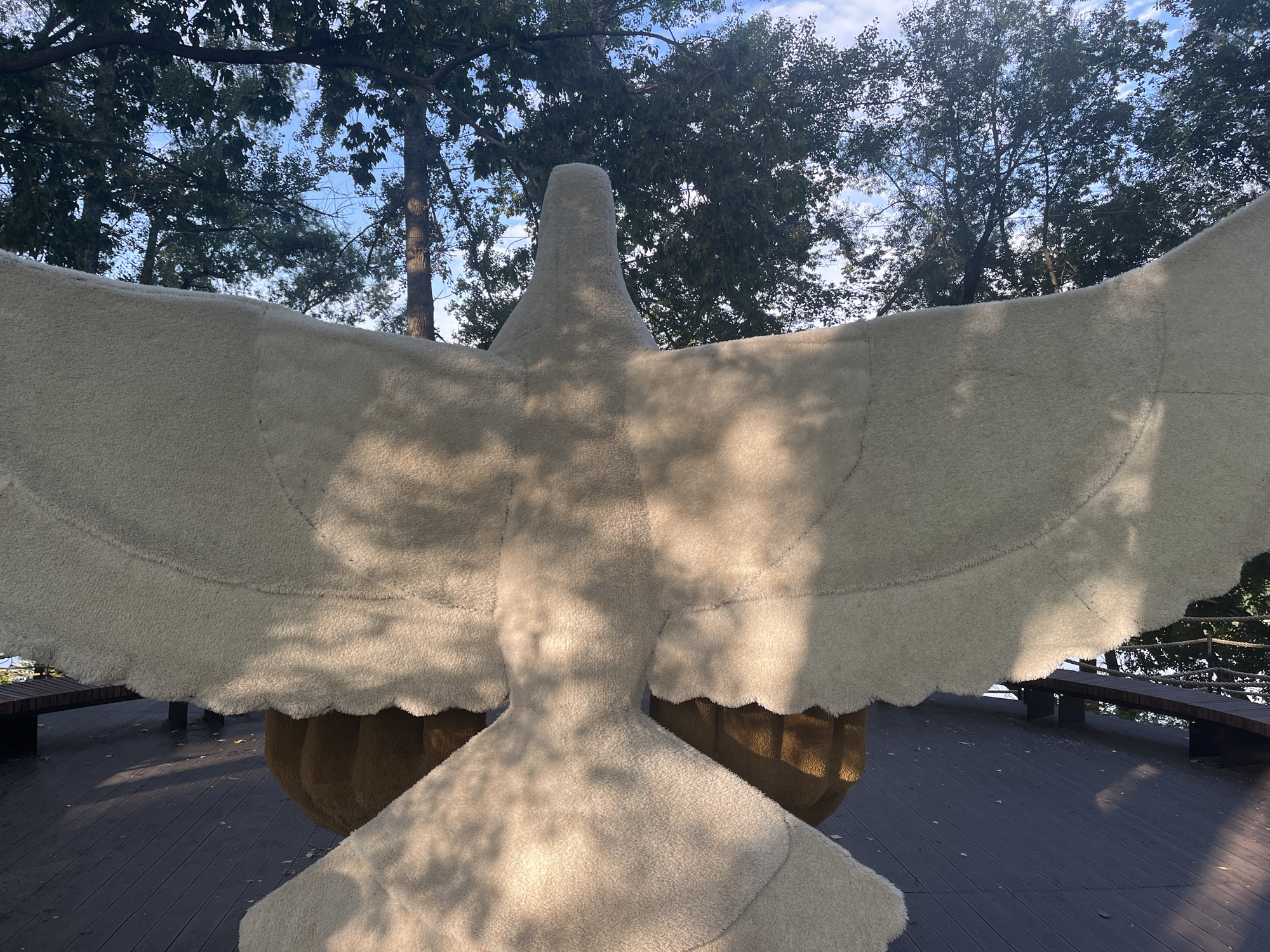 The image shows a large, fuzzy, light-colored sculpture of a bird with its wings spread wide, viewed from behind. The sculpture appears to be made of a plush material. It is positioned outdoors on a dark wooden deck. Trees with green foliage and a blue sky with scattered clouds are visible in the background. The sun casts dappled shadows from the trees onto the sculpture and the deck. To the left and right sides of the deck, benches are visible. This sculpture is located in the city of Blagoveshchensk, Russia, specifically in the city park named after the poet Alexander Pushkin, near the Amur River.