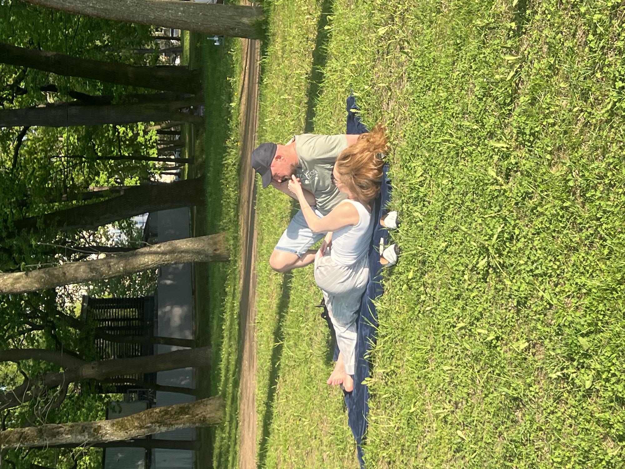 A medium, eye-level shot depicts a man and woman on a dark-colored blanket in a grassy area with trees along a pathway on a sunny day, seemingly in a park. The man, wearing a baseball cap, green shirt, and light-colored shorts, appears to be holding something small near his face, possibly a cigarette or electronic cigarette, while the woman, wearing a white top and light-colored pants, is positioned beside him and appears to be holding his hand. The background reveals a park-like setting with tall trees and a walking path to the left. The image was taken in the city of Saint Petersburg, Russia.
