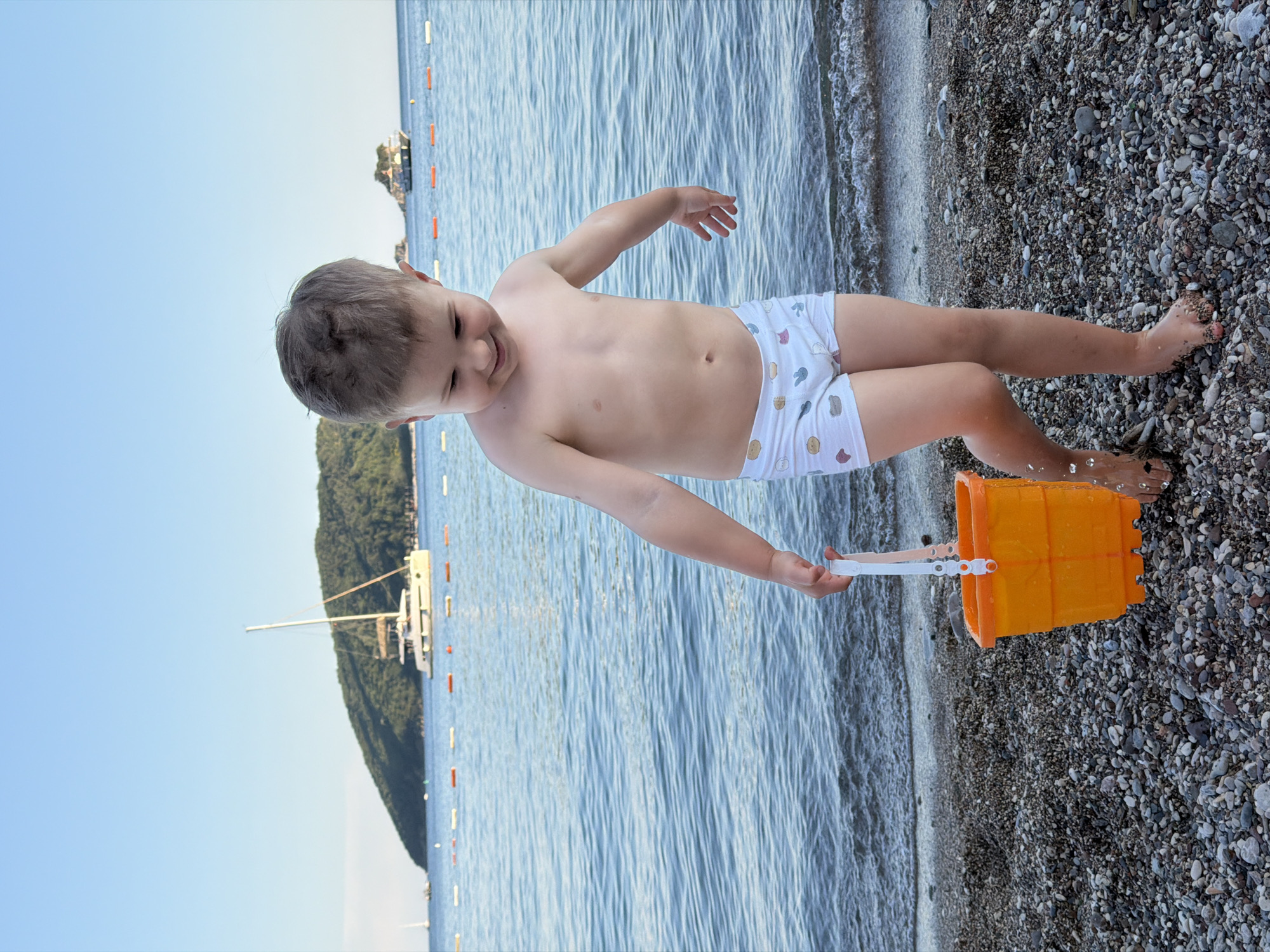 A young child wearing cartoon-patterned underwear stands on a pebble beach, holding an orange bucket. The child is near the shoreline of a blue body of water. Behind the water, there's a lush green hill with some visible structures, likely boats or buildings. The sky is light blue. This appears to be a beach scene, likely in Budva, Montenegro, given the landscape's characteristics. The child is interacting with the beach and water, presumably filling or emptying the bucket.