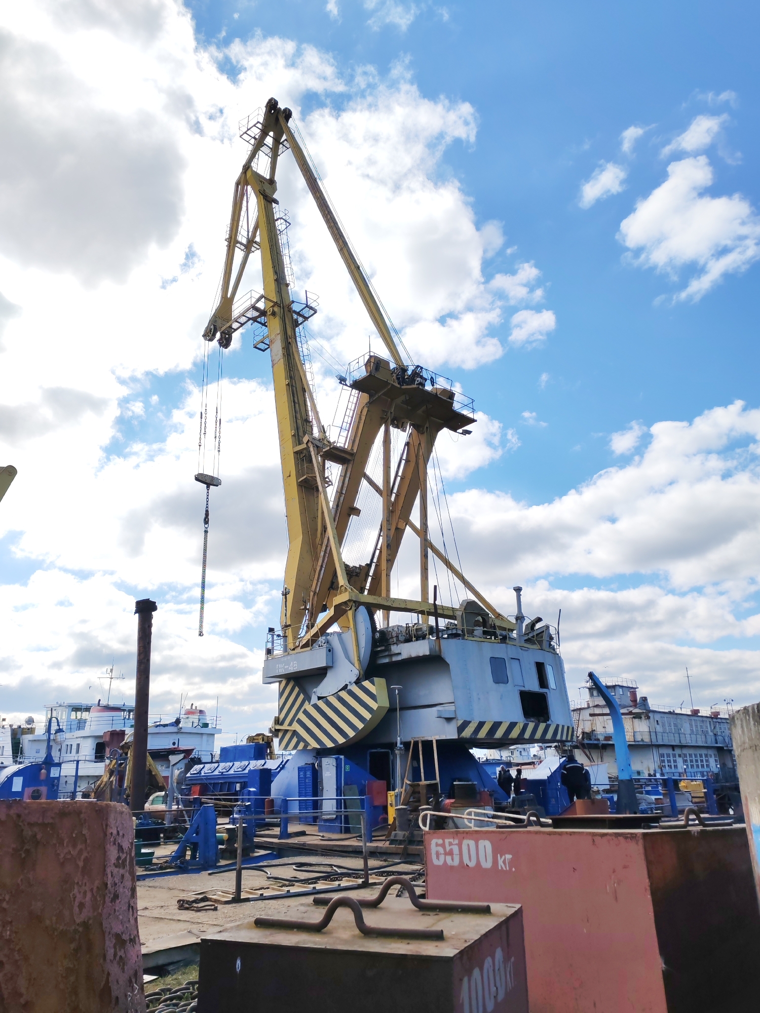 The image shows a large, yellow and gray port crane in Rybinsk, Russia. The crane has a capacity marking of "6500 кг" visible on a rusty metal structure in the foreground. In the background, there are several boats docked near the crane. The sky is blue with scattered white clouds. The ground appears to be a working area, likely a dock or shipyard, with various metal components and structures scattered around.
