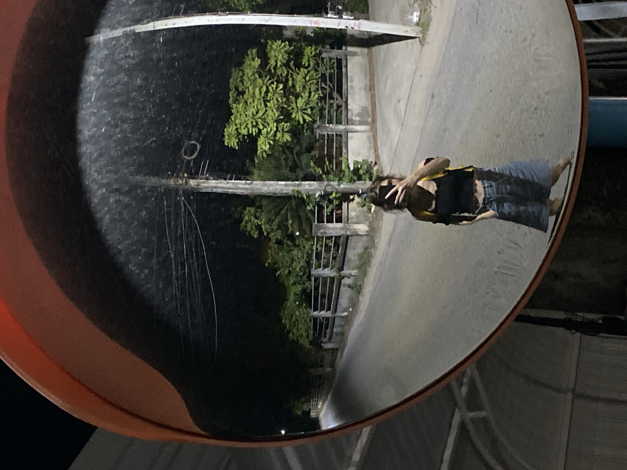 The image shows a convex mirror mounted on a post reflecting a scene in Baan Mae Nam, Thailand. In the reflection, a person wearing a black top and denim shorts is visible, standing on a paved surface. The background includes vegetation, a fence, utility poles with wires, and a dark night sky on the left side of the mirror. The mirror itself has an orange border.