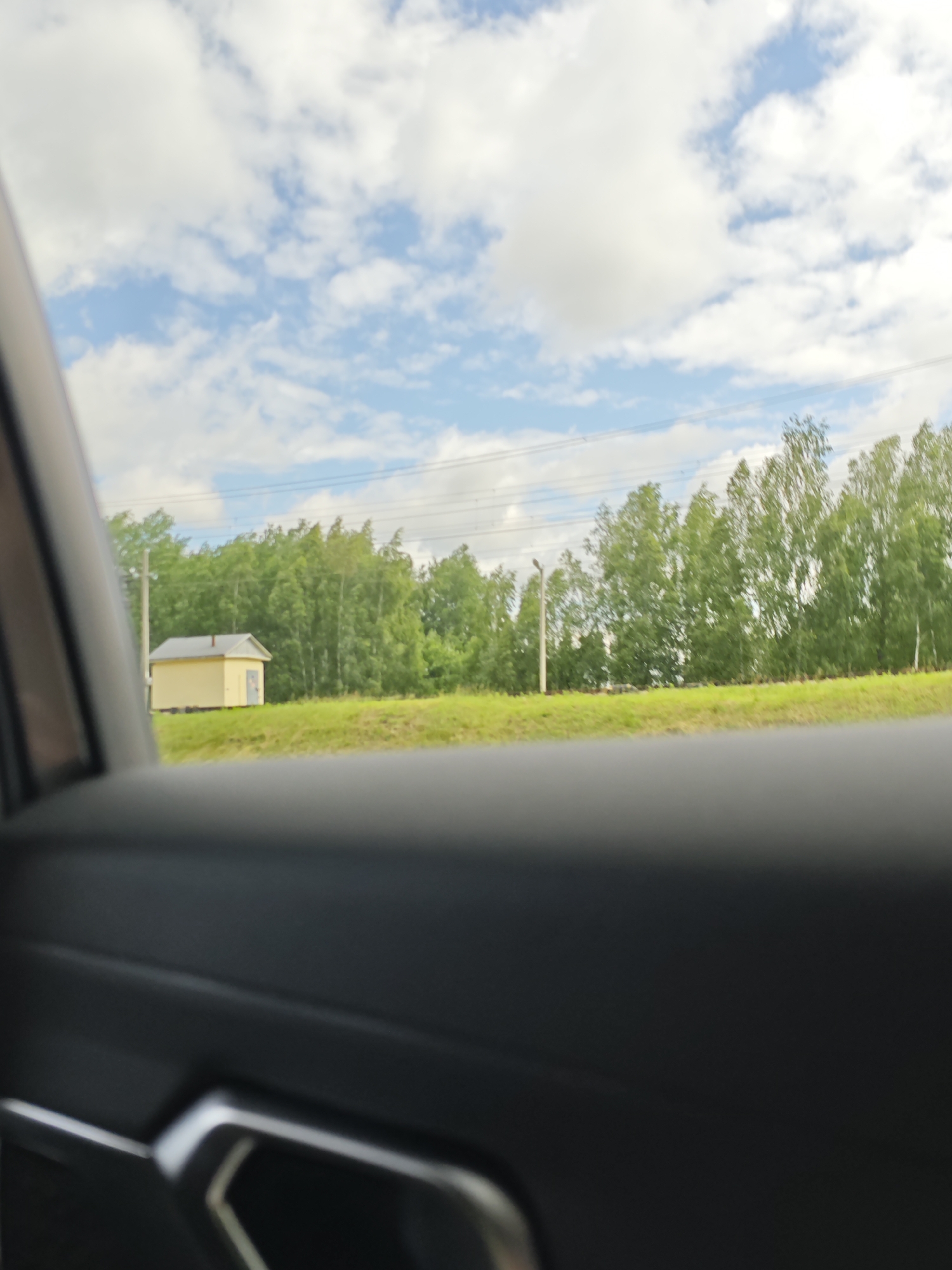The image captures a view from inside a vehicle, looking outwards through a partially open window or door. In the foreground, the dark interior of the car is visible, with a portion of a door handle or armrest in focus on the lower left. Beyond the car's interior, a bright, sunlit scene unfolds. A small, light yellow rectangular building with a grey roof and a dark door is situated in the middle ground, to the left of the frame. It sits on a grassy slope. Behind the building, a dense line of green trees stretches across the horizon, with some power lines visible above them. The sky is a mix of blue with fluffy white clouds. The overall impression is of a rural or suburban landscape viewed during the daytime.