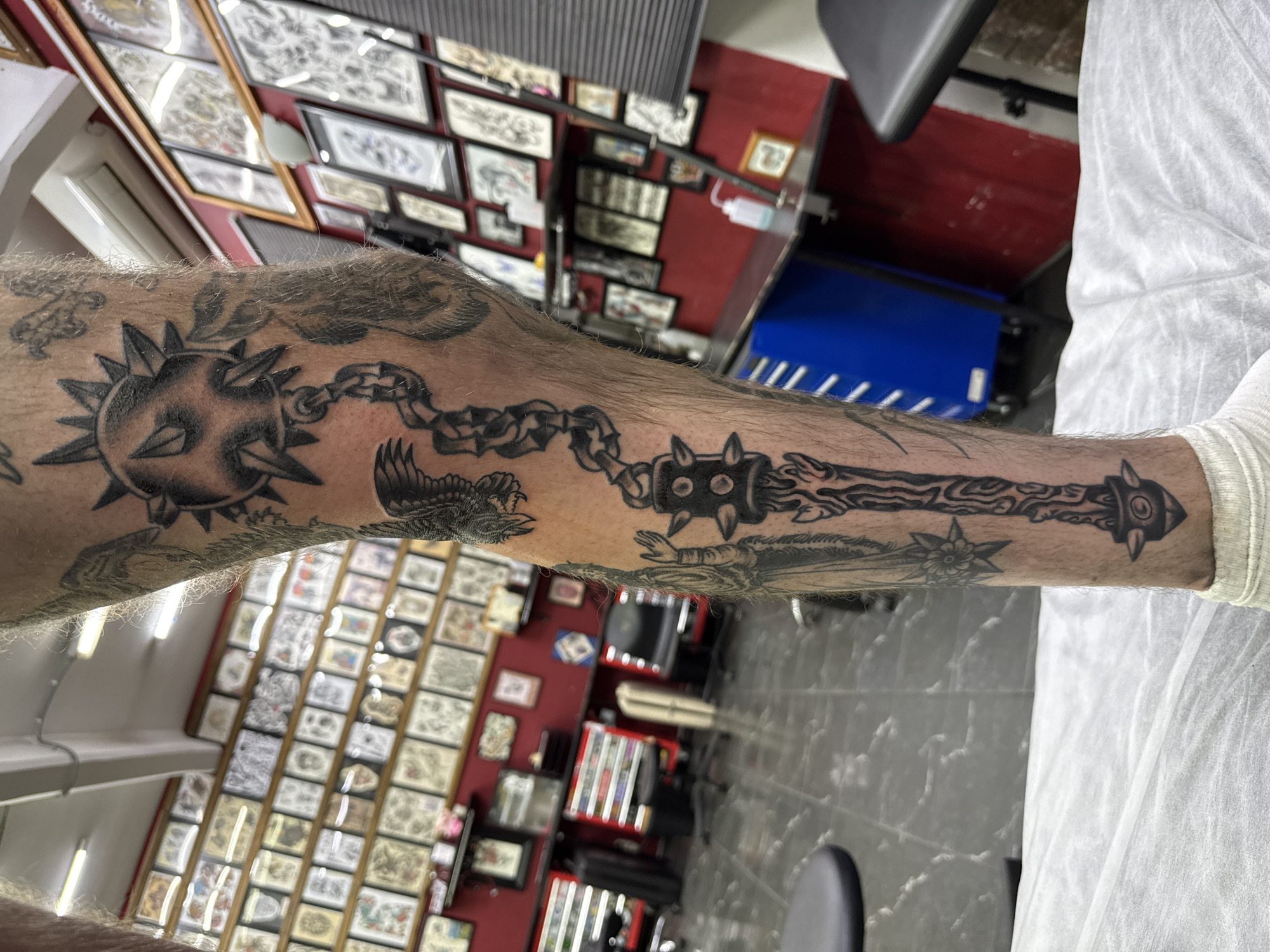 The image depicts a person's arm with a tattoo of two spiked maces connected by a chain, with other tattoos nearby, set against the background of a tattoo studio. The tattoos are in black ink. The studio has framed tattoo designs on the wall, along with tattoo equipment and chairs. The image provides no specific location in Moscow, Russia.