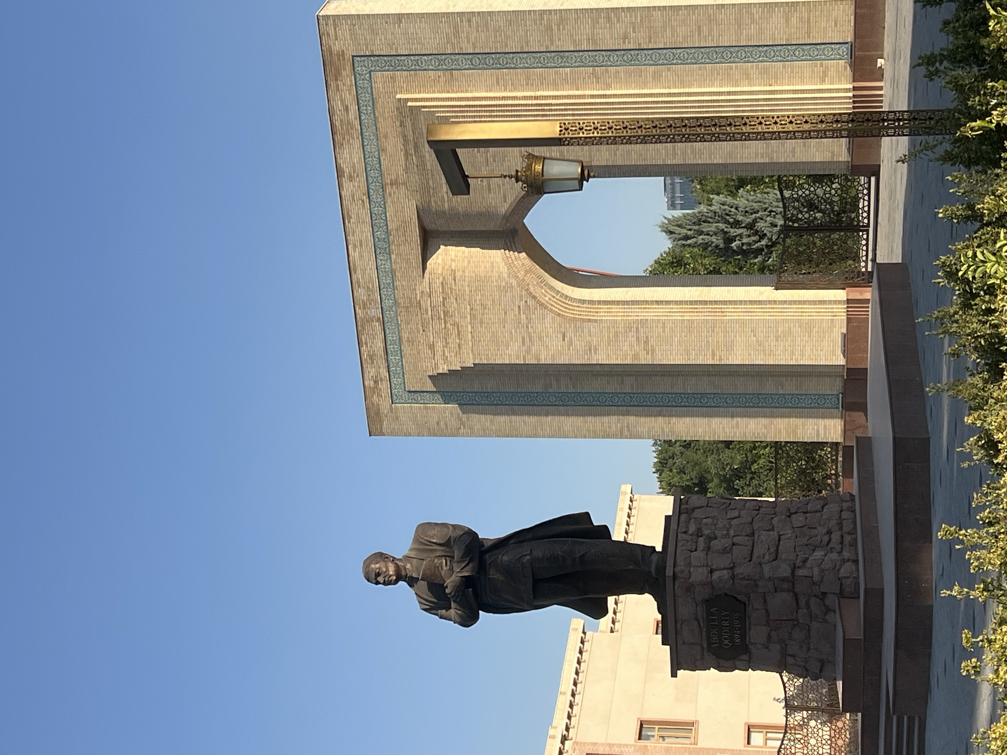 This media file features a bronze statue of a man in formal attire, possibly a historical figure or a statesman, standing on a stone pedestal. The statue is positioned in an outdoor setting with a clear blue sky in the background. To the left and above the statue is a section of a building constructed from light-colored bricks, adorned with decorative blue tiles and gold accents. A traditional-looking lantern hangs from an ornate metal fixture. The building's architectural style suggests a public space or monument.
To the right of the statue, lush green foliage frames the scene, hinting at a well-maintained park or garden. The ground is paved with stone tiles, and a dark stone structure is visible near the base of the statue. The overall impression is one of solemnity and historical significance.
A plaque on the pedestal of the statue displays visible text that reads "ABDULLA QODIRIY 1894-1934". This indicates the statue is of Abdulla Qodiri, an important figure in Uzbek literature and history. The time of day appears to be morning or late afternoon, judging by the clear, bright sunlight casting shadows. The weather seems to be fair and sunny. The setting is likely a public square or monument in Tashkent, Uzbekistan, given the provided location context and the architectural style.