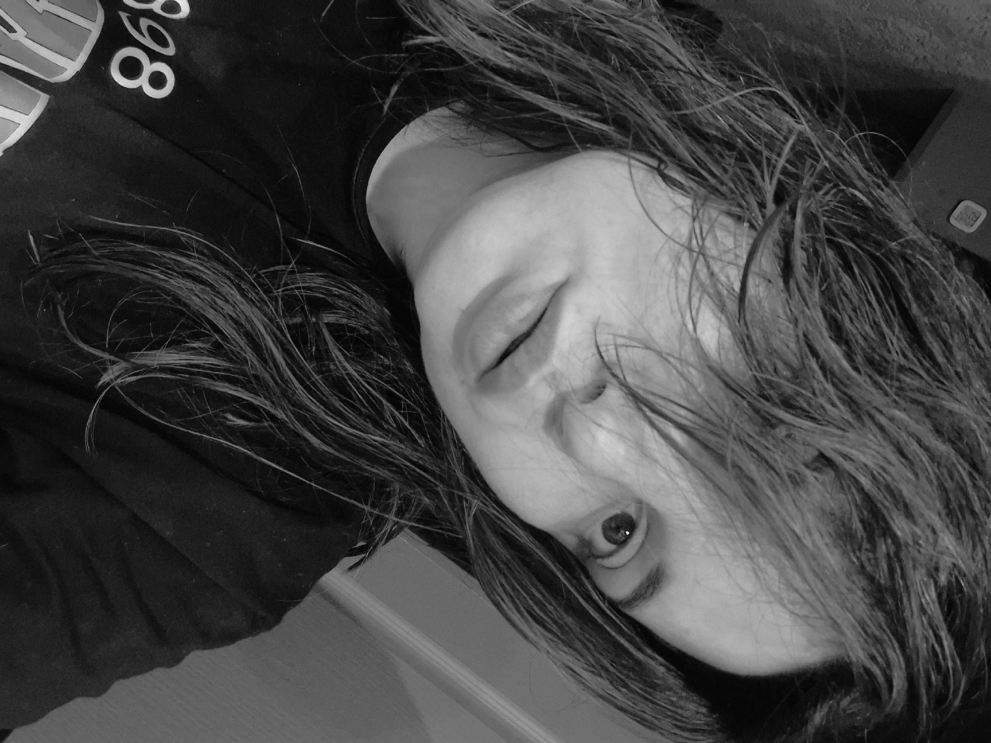 The image is a black and white close-up portrait of a single individual, appearing to be a young adult female. Her head is tilted to the viewer's left, and her gaze is directed towards the camera. Her right eye is clearly visible, while her left eye is partially obscured by strands of dark, seemingly damp or wet hair that fall across her forehead and face. Her lips are slightly parted. She is wearing a dark, possibly black, crew-neck shirt or sweatshirt, on which the partial white numerals "898" are visible on the lower left. The background is dark and indistinct, suggesting an indoor environment. In the upper left portion of the background, a small, light-colored square object, resembling a thermostat or a wall-mounted sensor, is present. The overall image has a grainy texture.