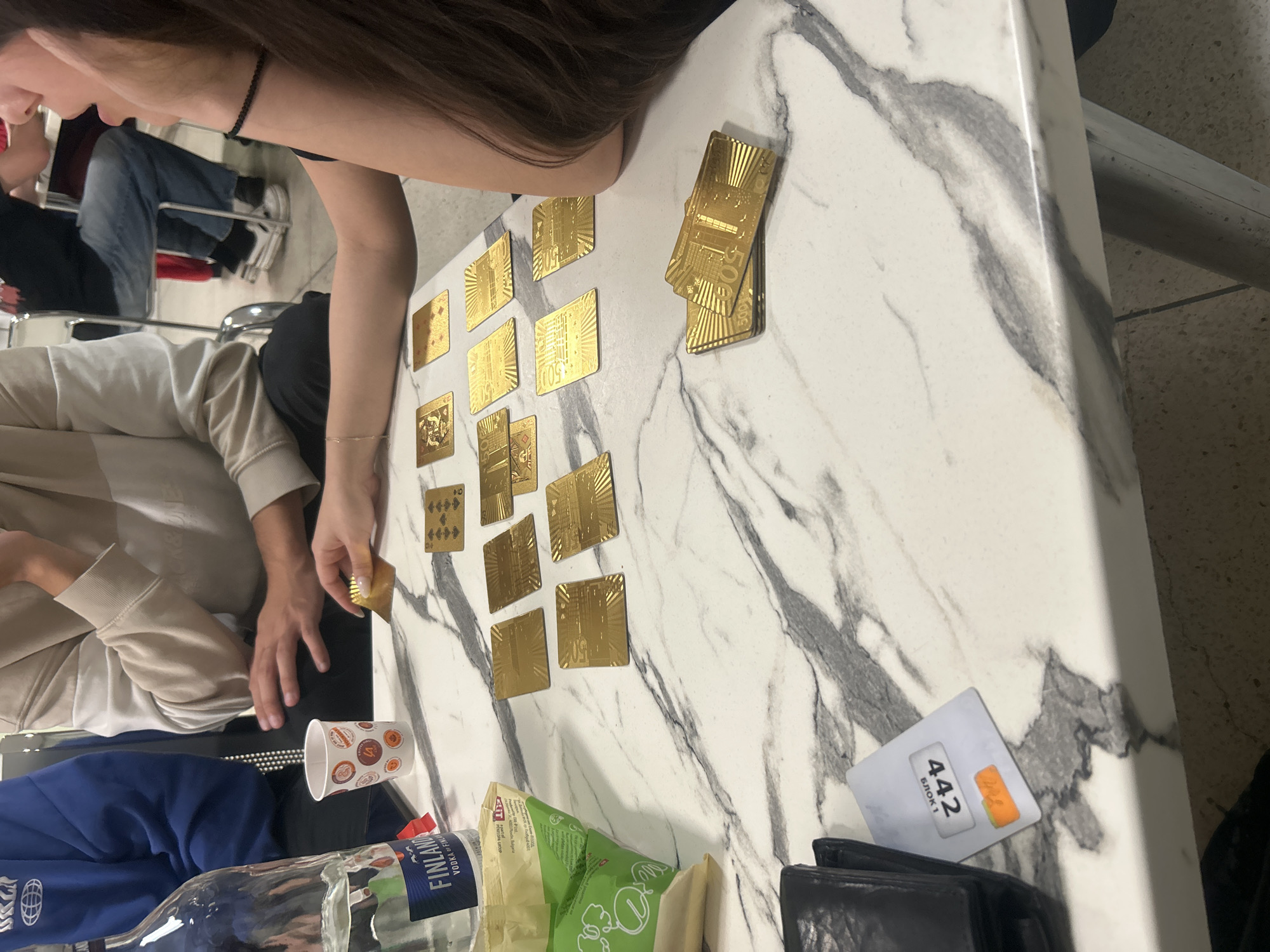 The scene depicts a group of people sitting at a table covered with a marble-patterned surface. Scattered across the table are gold-colored playing cards. A bottle of Finlandia Vodka and a bag of snacks are visible near the edge of the table. A white card with the number "442 БЛОК 1" and an orange sticker is present. The location is Veliko Tarnovo, Bulgaria.