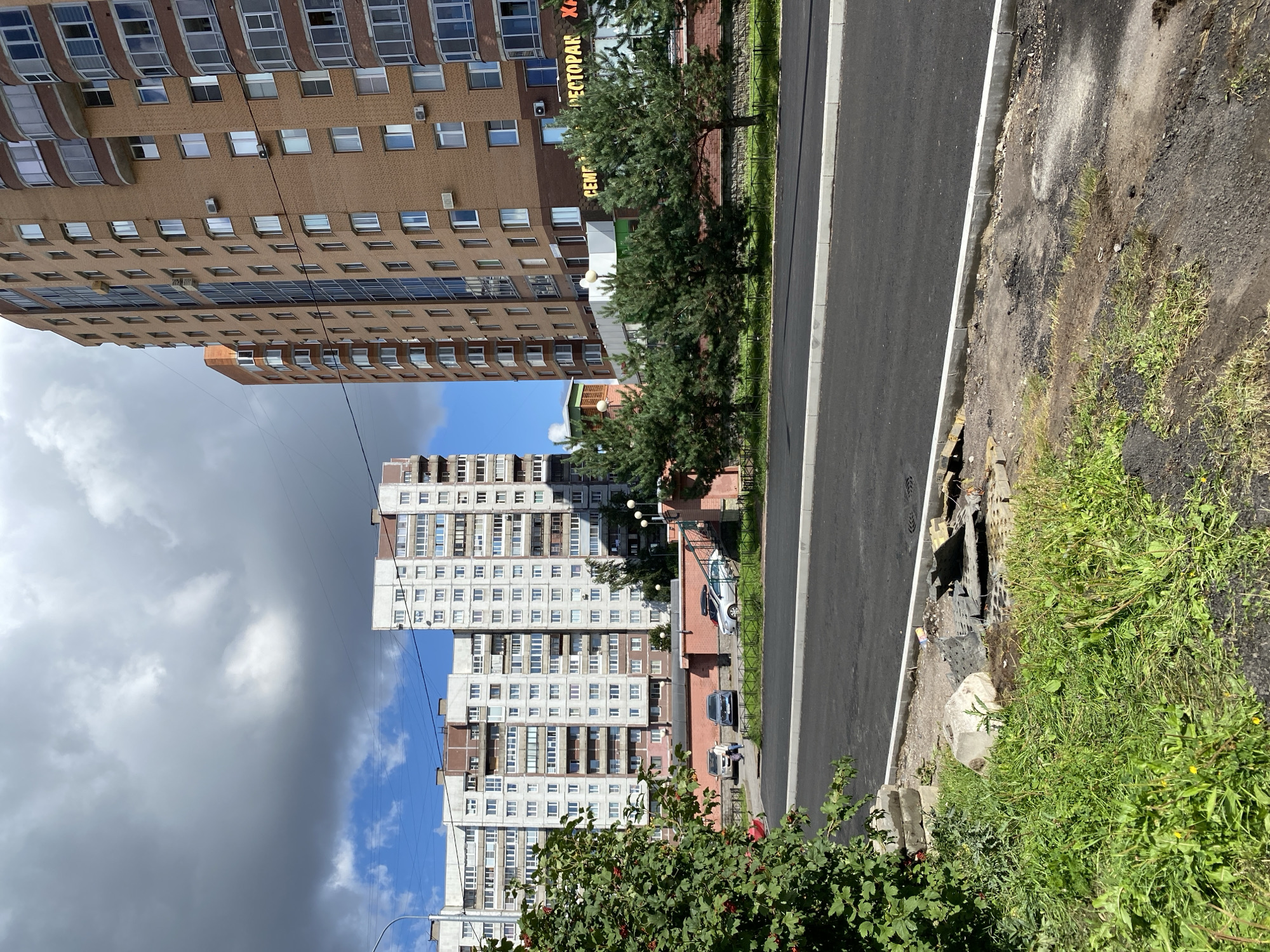 The image depicts a street scene in Saint Petersburg, Russia, under a partly cloudy sky. In the foreground, a newly paved asphalt road runs horizontally, bordered by a curb and a patch of overgrown grass and debris on the left. The debris includes concrete blocks, broken asphalt, and a piece of wood. To the right of the road, a row of evergreen trees is visible in front of a low brick wall. Beyond the trees, a multi-story building with a facade of brown brick and multiple windows dominates the right side of the frame. Several windows on this building are open, and air conditioning units are mounted on the facade. Above the trees and brick wall, a sign reading "СЕМЕЙНЫЙ РЕСТОРАН" (Family Restaurant) is partially visible.
In the background, on the left side of the image, a large, multi-story apartment building with a white and grey facade and numerous windows is visible. Further in the distance, another tall apartment building with a similar design is seen. Parked cars, including a white sedan and a dark grey sedan, are on a paved area between the apartment buildings. A person is walking near these cars. The overall environment suggests a residential urban area.