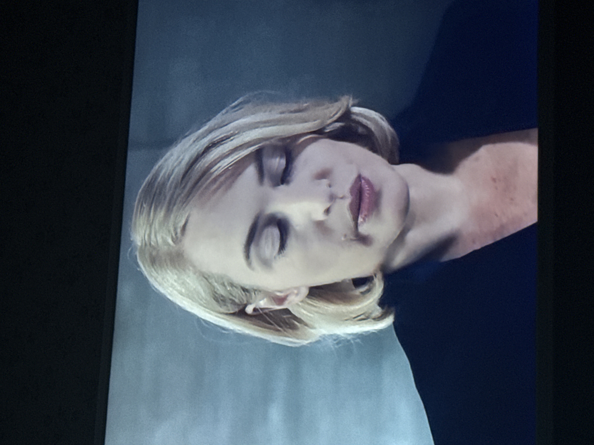 A medium shot of a woman with blonde hair and closed eyes, lying down against a light blue or grey background. She is wearing a dark top. Her face is tilted slightly upwards. There is no indication that this image was taken in Mytishchi, Russia.