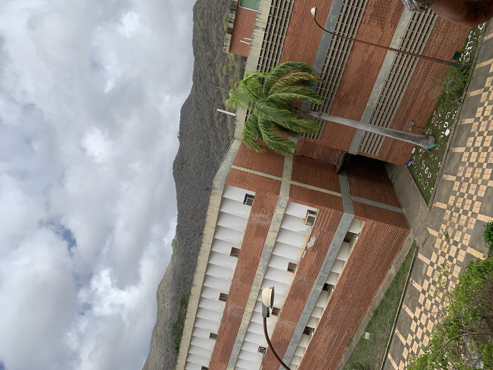 The image shows a multi-story building with red brick walls and white vertical accents. The building is constructed on a hillside, with a mountainous, tree-covered landscape in the background. A palm tree stands in front of the building, leaning towards the left. The ground in front of the building features a checkered pavement pattern. There are several windows on the building, some of which appear to have air conditioning units. A street lamp is visible to the right of the palm tree. The sky is overcast with clouds. The scene is located in Las Piedras, Venezuela.